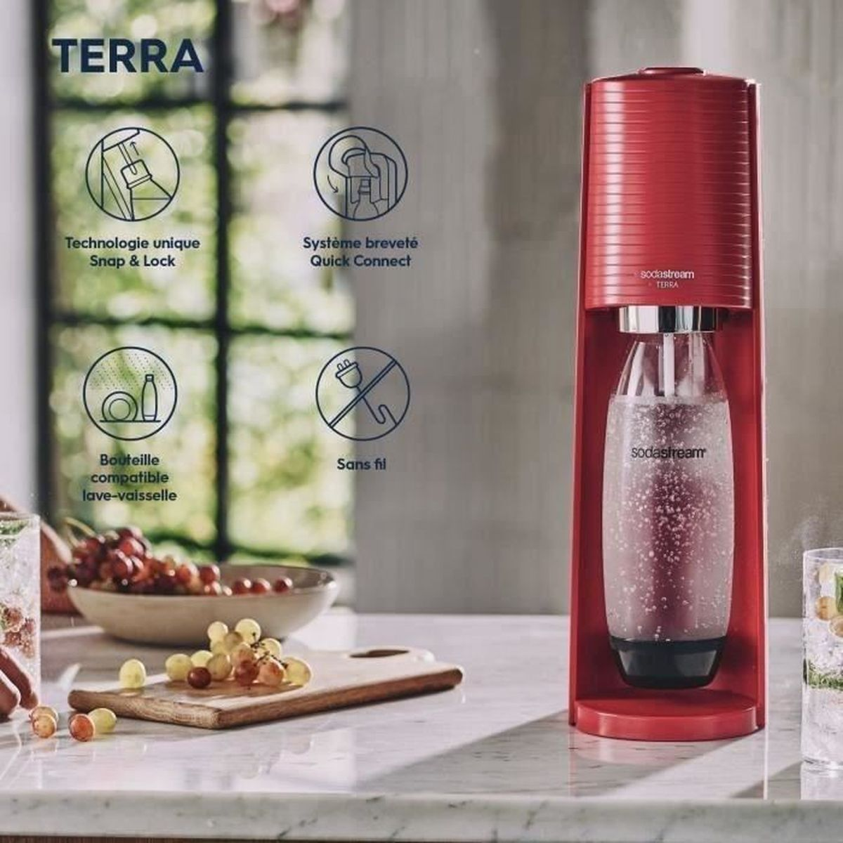 Rote SodaStream-Maschine mit Glasflasche auf Marmoroberfläche. Trauben und ein Schneidebrett im Vordergrund.