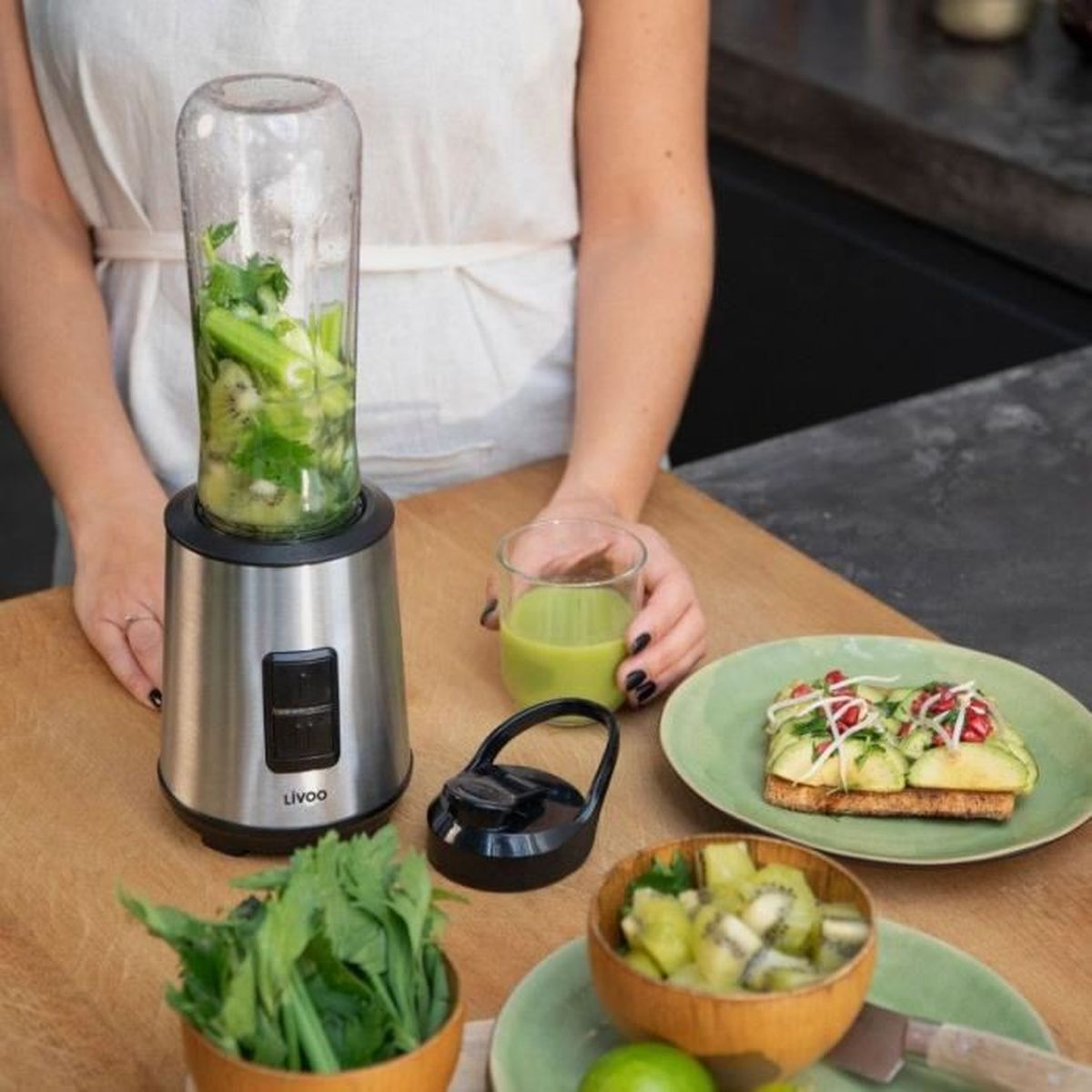 Eine Person bereitet einen Smoothie zu. Ein Mixer, ein Glas Saft und ein Teller mit Essen stehen auf einem Holztisch. Obst und Gemüse sind vorhanden.