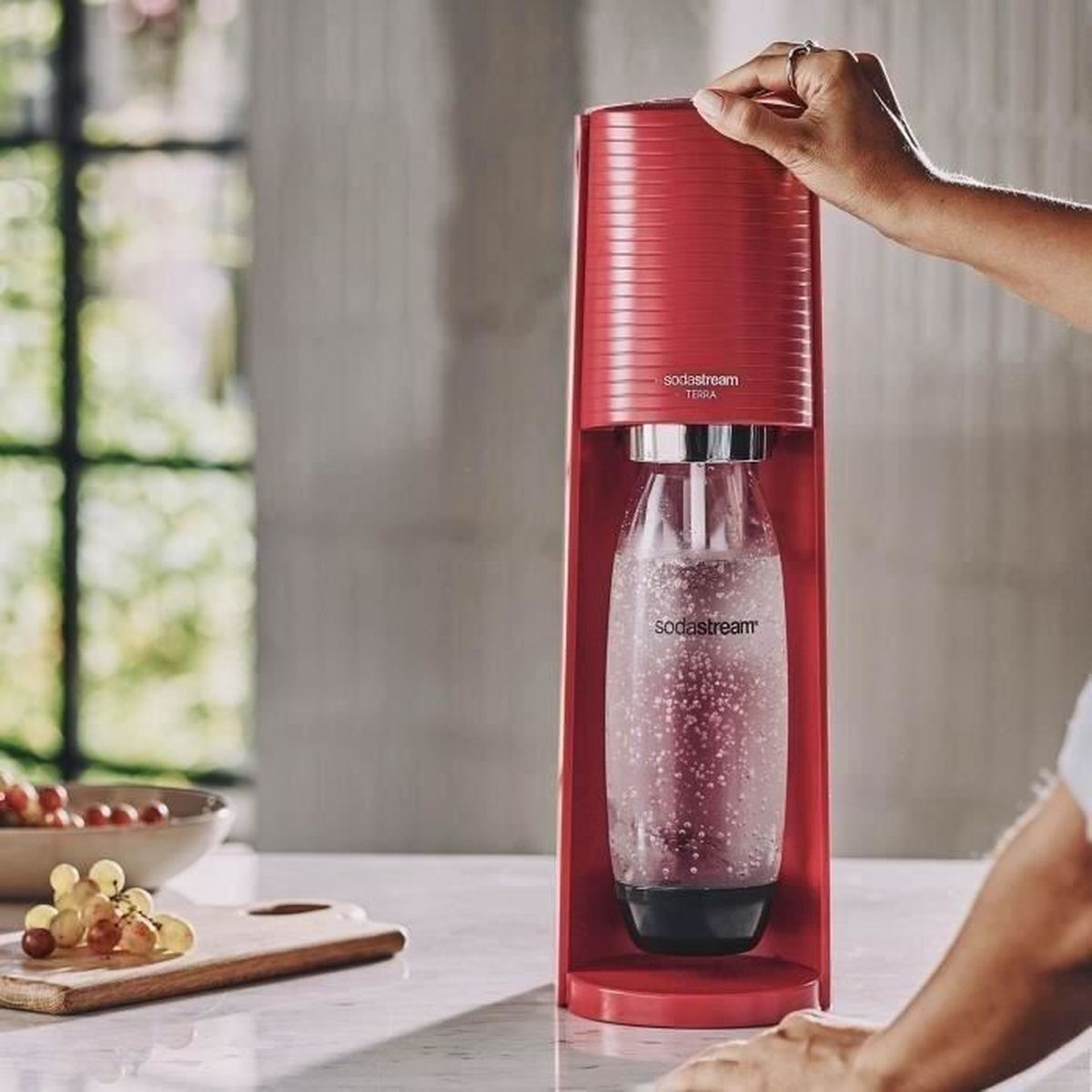 Ein rotes SodaStream-Gerät mit einer durchsichtigen Flasche. Eine Hand bedient es, Trauben und ein Schneidebrett liegen auf dem Tisch.