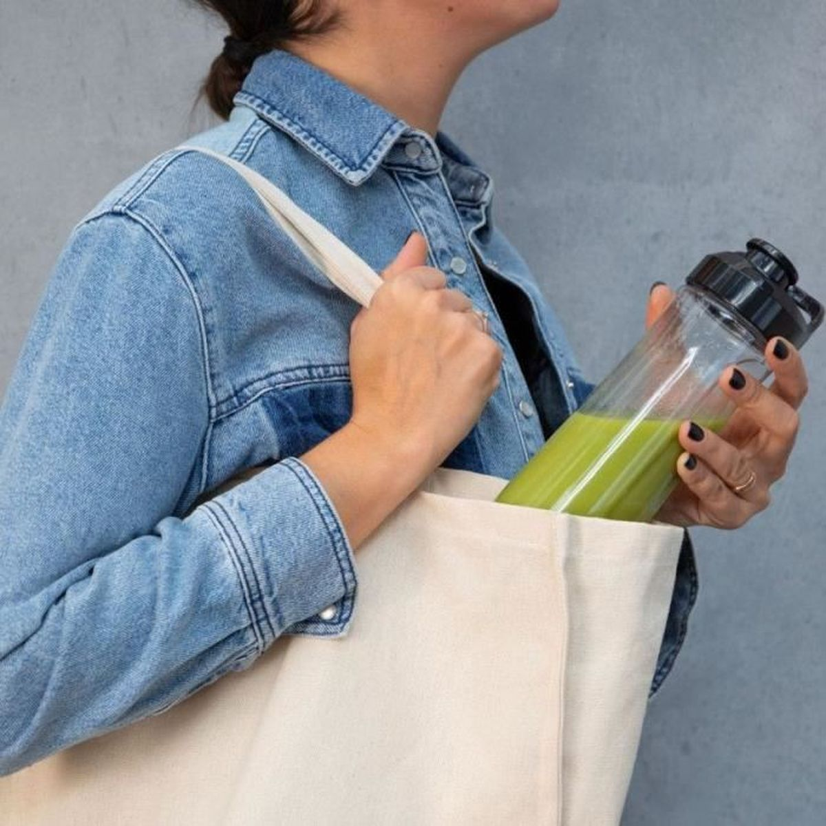 Eine Frau in einer Jeansjacke hält eine Einkaufstasche mit einer grünen Smoothie-Flasche. Schwarzer Nagellack und Ringe sind sichtbar.