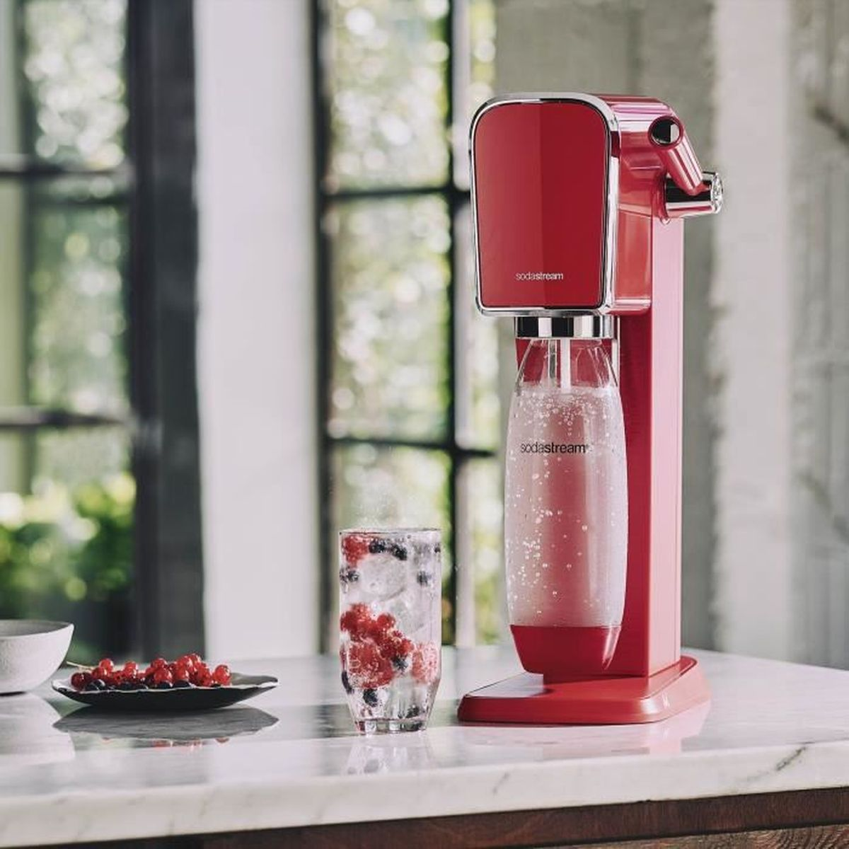 Czerwone urządzenie SodaStream stoi obok szklanki wody z jagodami i lodem. Talerz z jagodami jest z przodu.