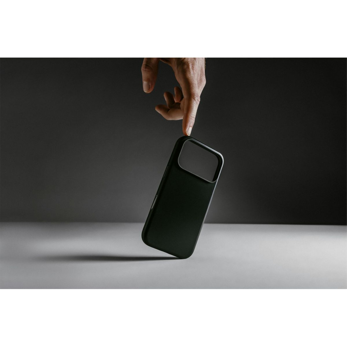 Una mano tiene una custodia nera per telefono sopra una superficie grigio chiaro. Sfondo grigio scuro. La custodia ha un ritaglio per la fotocamera.