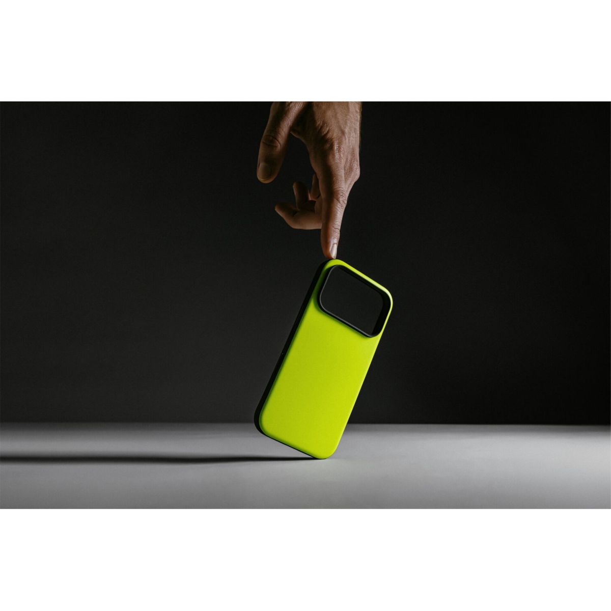 Una mano tocca una custodia per telefono verde lime con un ritaglio nero per la fotocamera. Lo sfondo è grigio scuro, con una superficie bianca sotto.