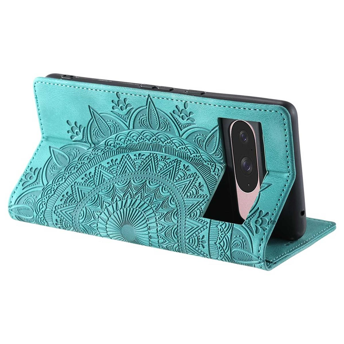 Eine türkisfarbene Handyhülle mit geprägtem Mandala-Design, die ein schwarzes Smartphone hält. Die Hülle ist offen und steht auf einer weißen Oberfläche.