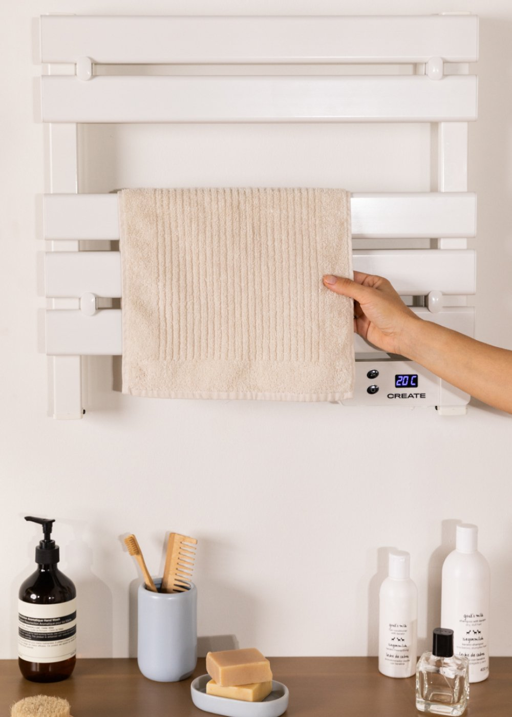 Una mano coloca una toalla beige en un toallero calefactado blanco. Debajo hay accesorios de baño, como jabón y artículos de tocador.