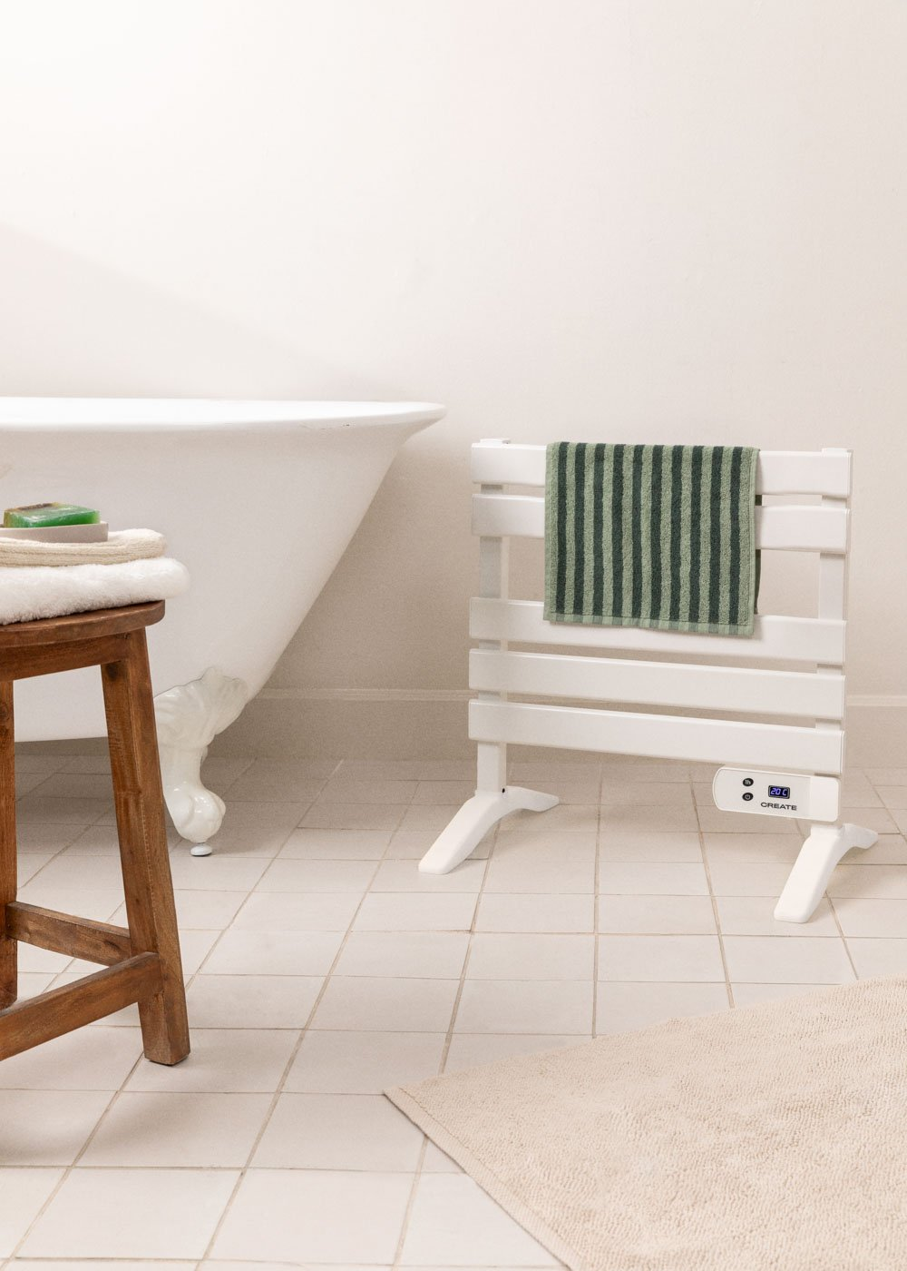 Escena de baño: toallero blanco con toalla verde. Bañera y taburete de madera con toallas y jabón. Alfombra beige sobre suelo embaldosado.