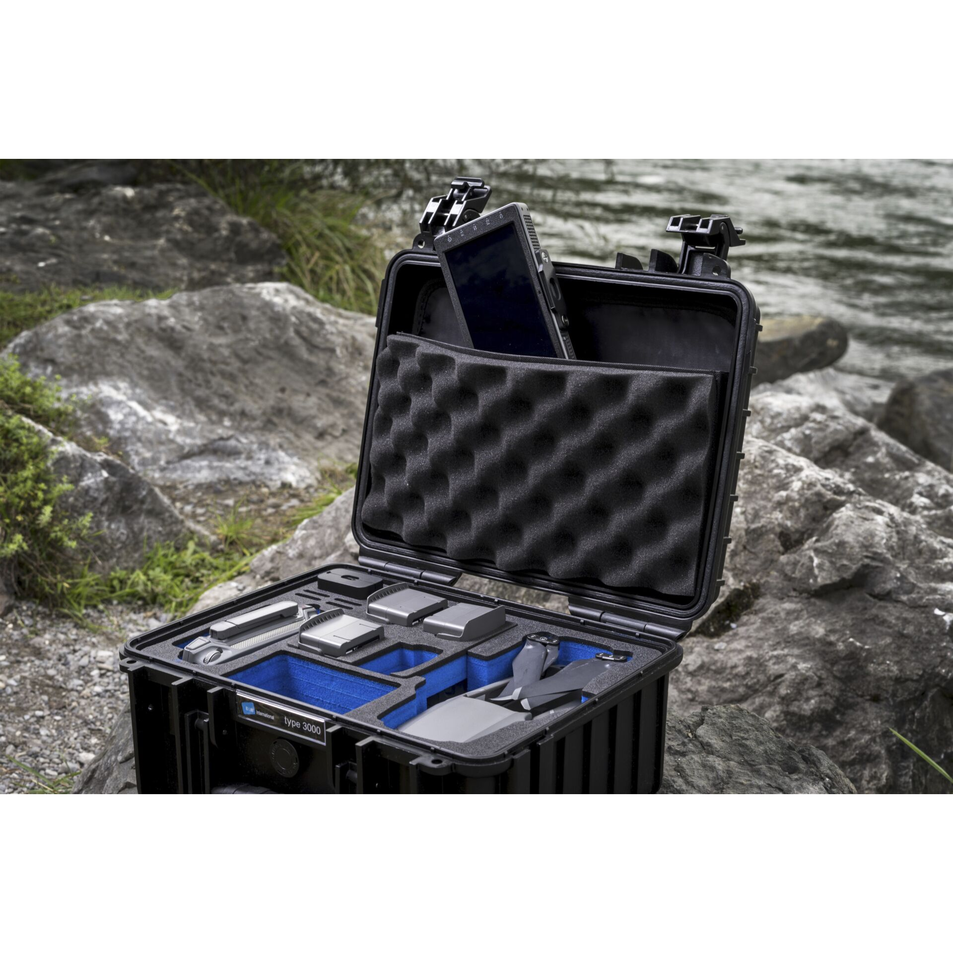 Schwarzer Koffer, geöffnet auf Felsen, zeigt Drohnenteile, Batterien und ein Tablet. Hintergrund: Wasser und mehr Felsen.