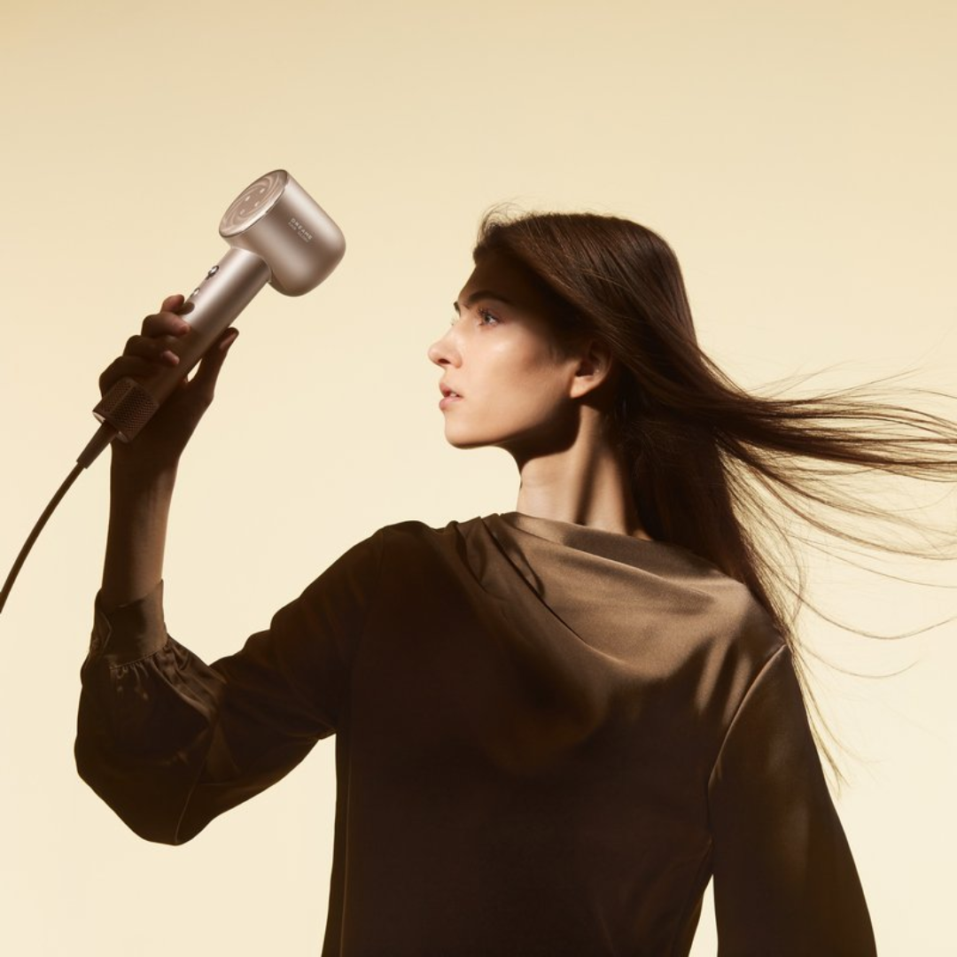 Una mujer sostiene un secador de pelo plateado, el pelo ondeando. Viste un top marrón contra un fondo crema. La escena está iluminada con luz suave.