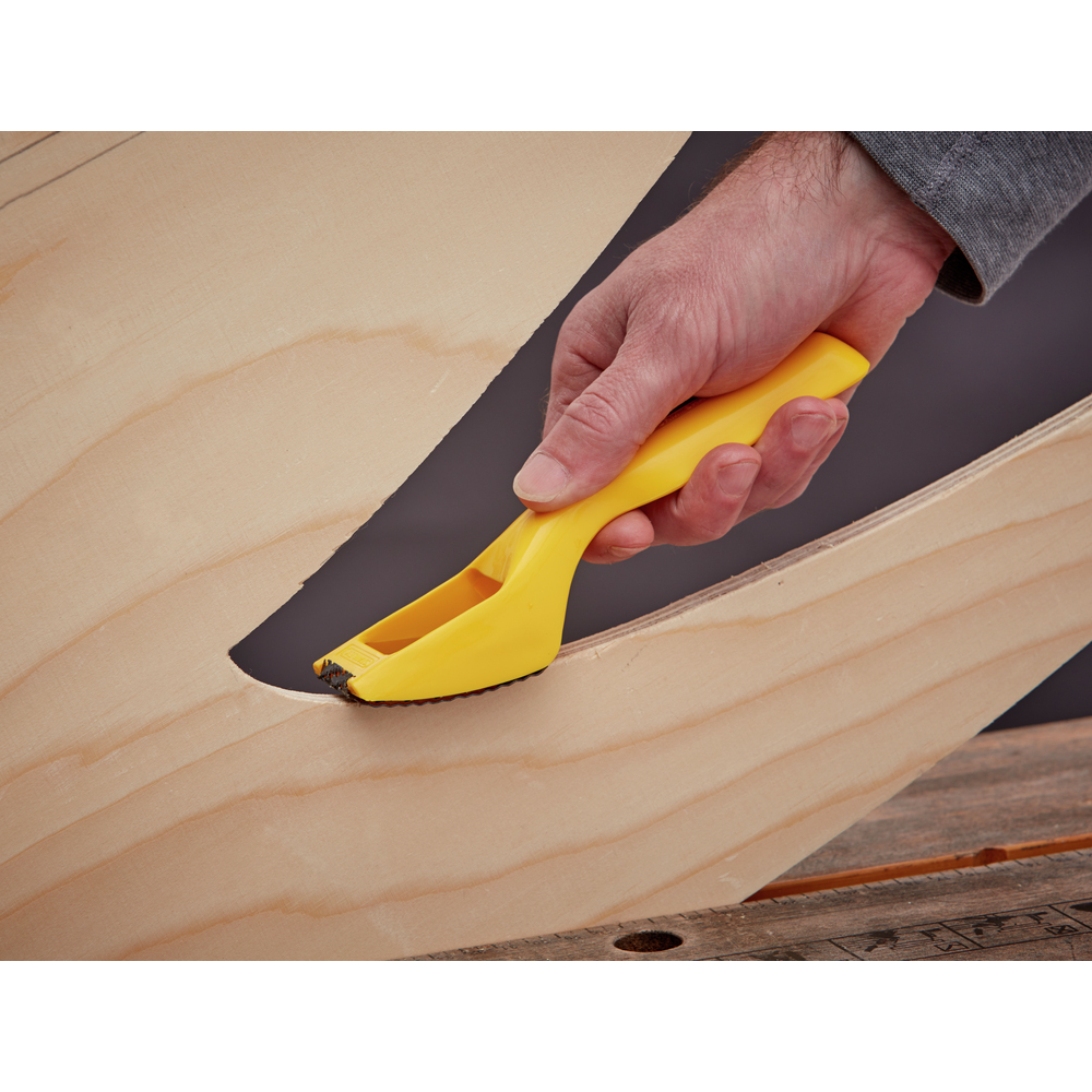 Eine Hand hält ein gelbes Werkzeug, um die Kante einer Holzplatte zu glätten. Die Platte hat einen dunklen, gebogenen Schnitt.