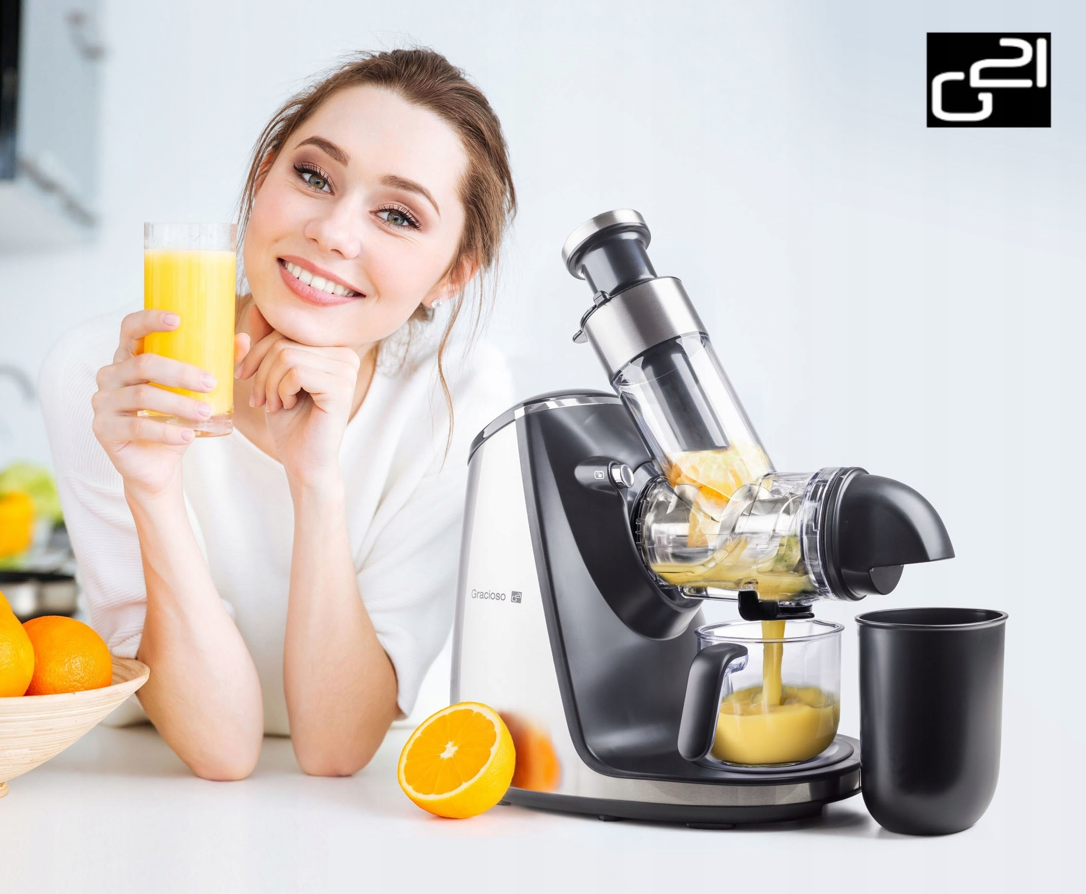 Uśmiechnięta kobieta trzymająca sok pomarańczowy. Sokowirówka z pomarańczami. Sok wlewa się do szklanki. Połówka pomarańczy na blacie.