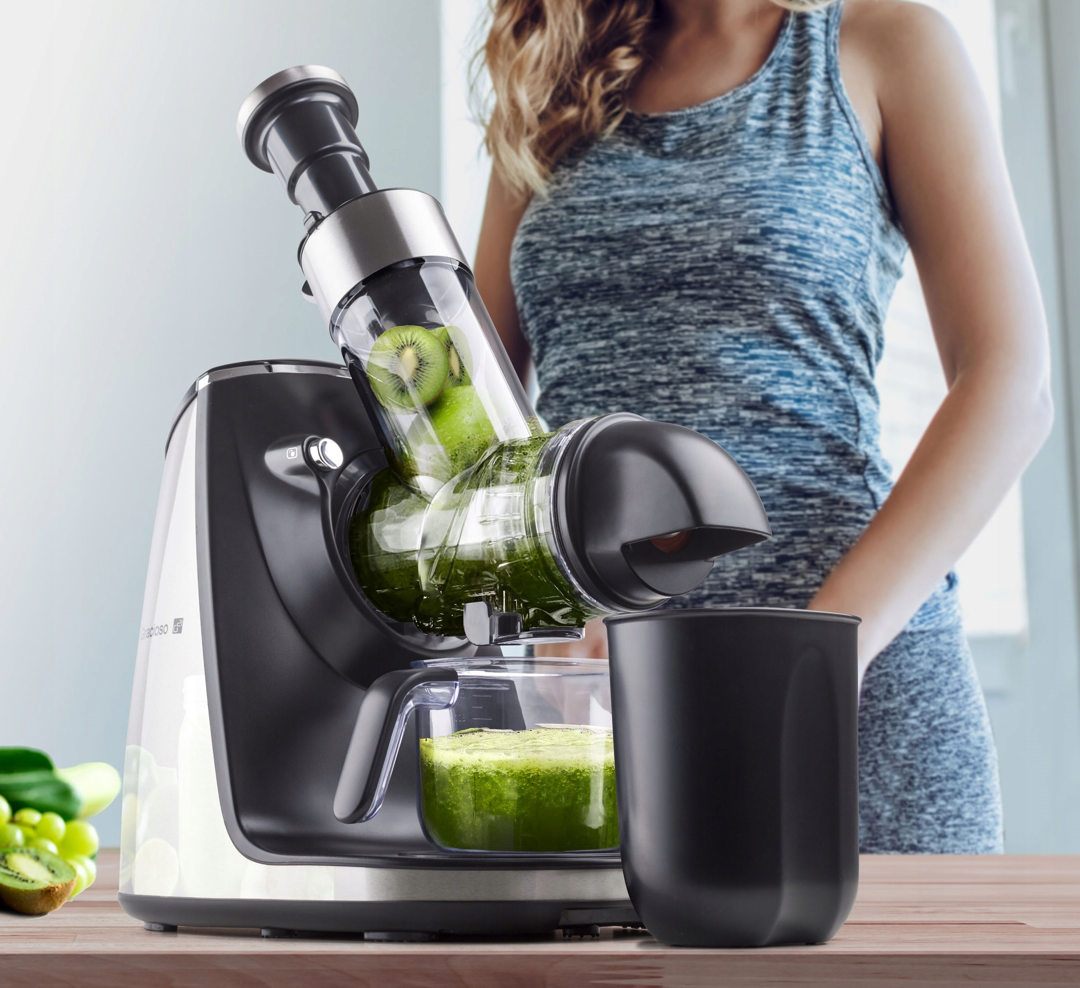 Kobieta przy wyciskarce do soku z kiwi. Wyciskarka ma zielony sok i kiwi w środku. Czarny kubek obok wyciskarki na drewnianej powierzchni.