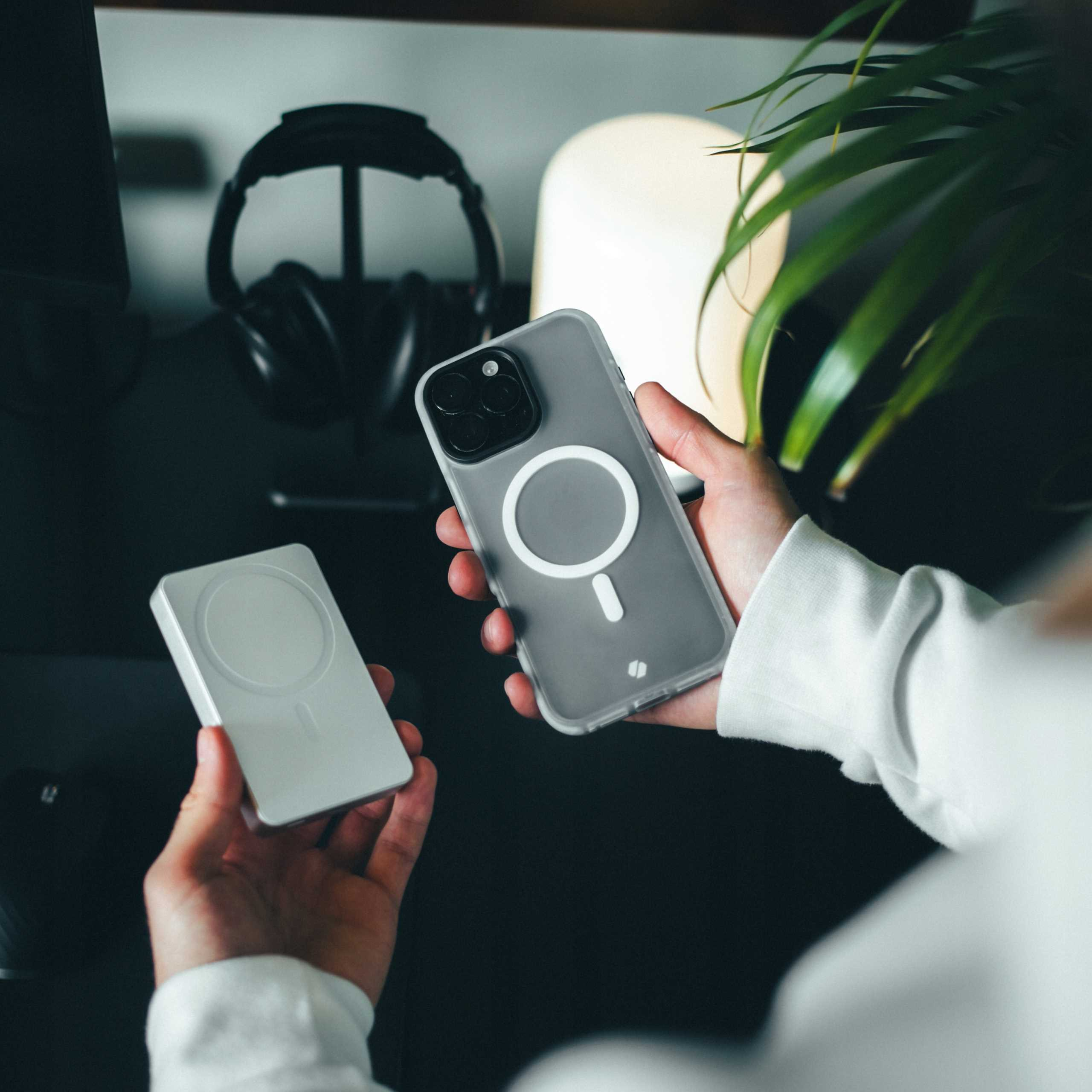 Ręce trzymające telefon w przezroczystym etui i białą podkładkę ładującą. Słuchawki i lampa widoczne w tle.