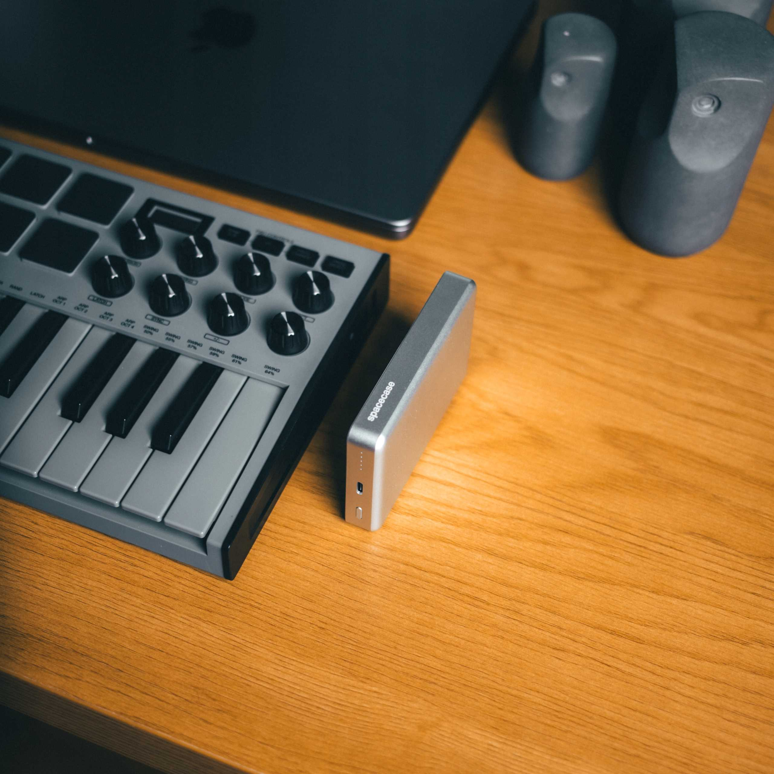 Szara klawiatura MIDI, srebrny power bank i laptop leżą na drewnianej powierzchni.