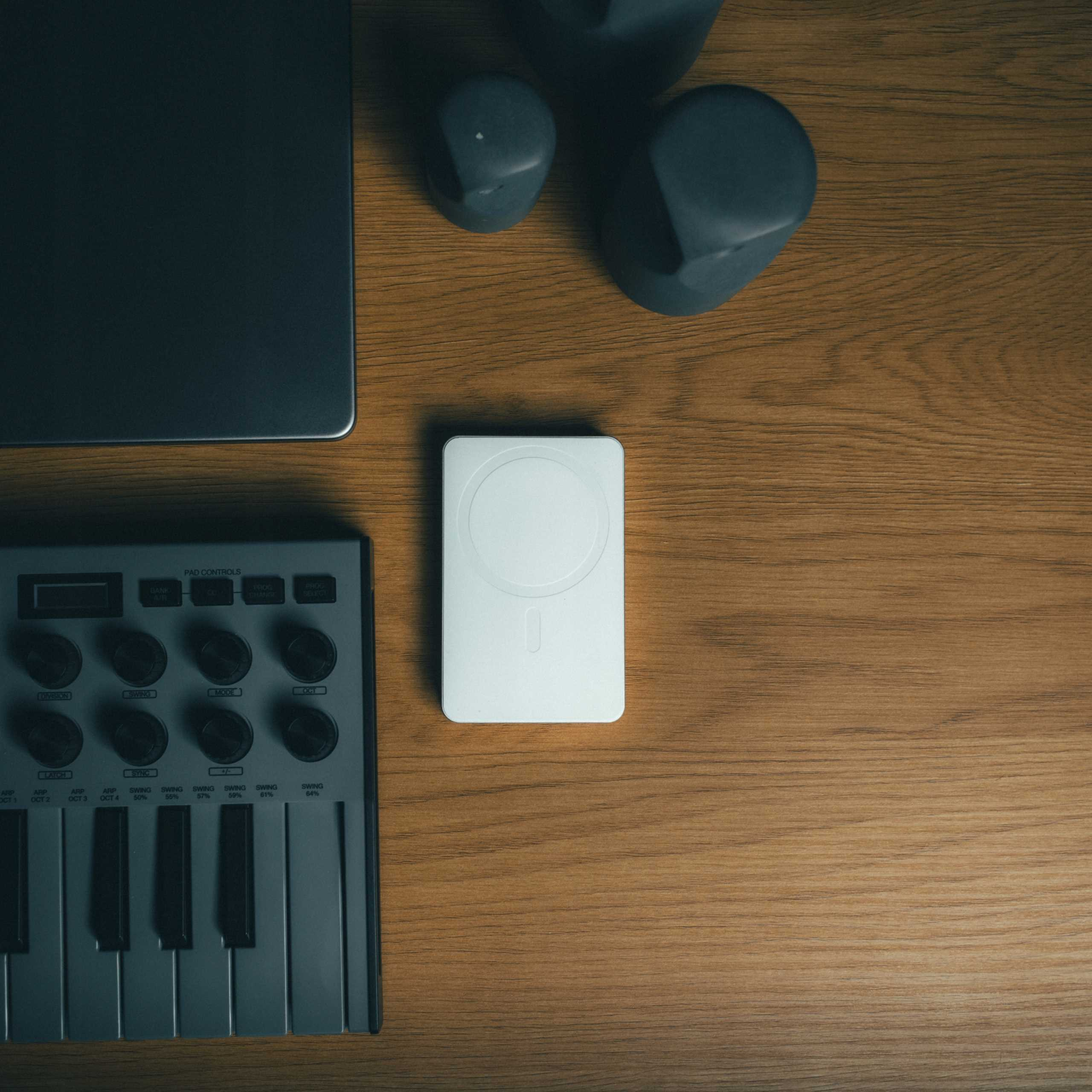 Drewniany stół z laptopem, białą podkładką ładującą, szarym syntezatorem i trzema ciemnoszarymi przedmiotami.