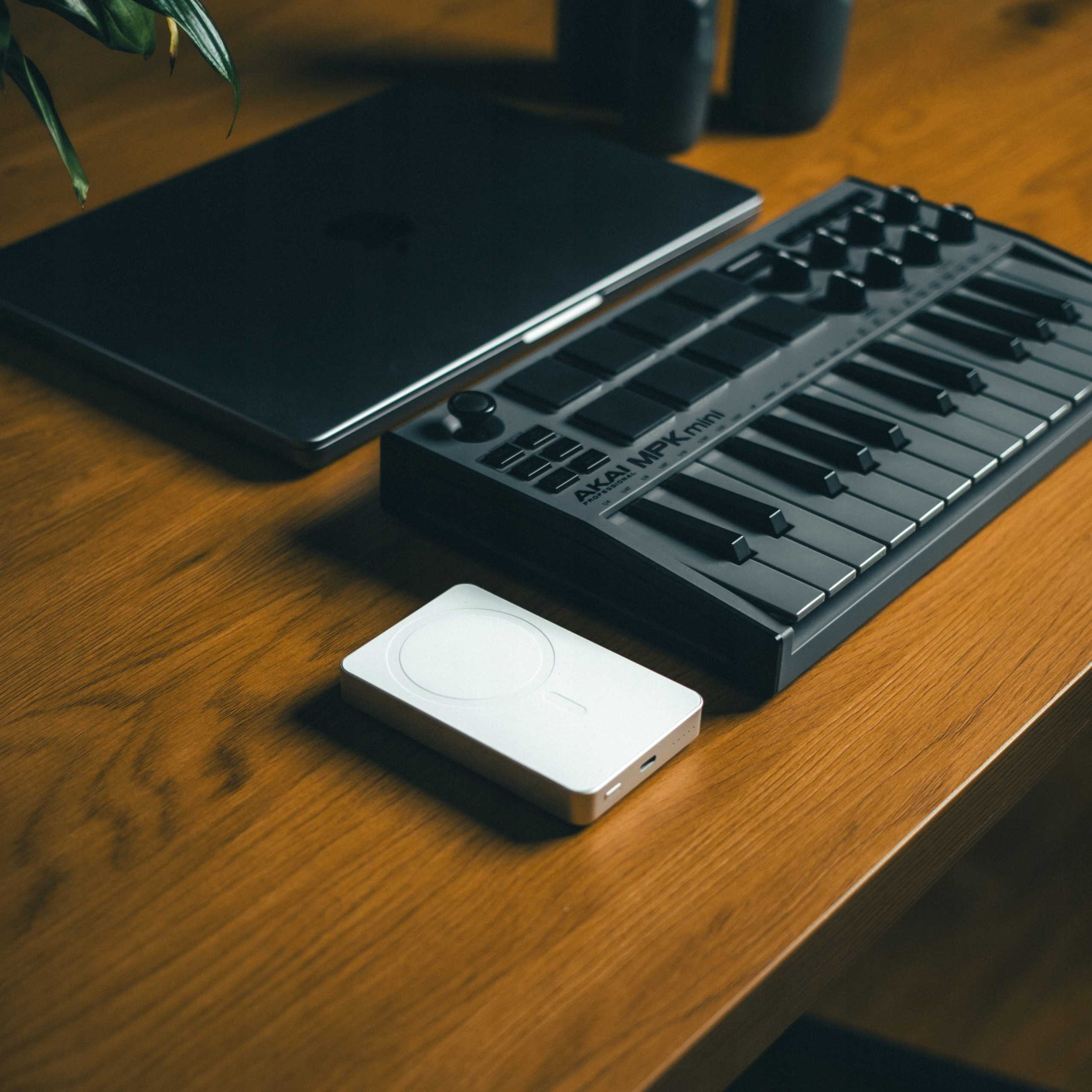 Laptop, klawiatura MIDI i białe urządzenie na drewnianym biurku. Roślina w lewym górnym rogu.