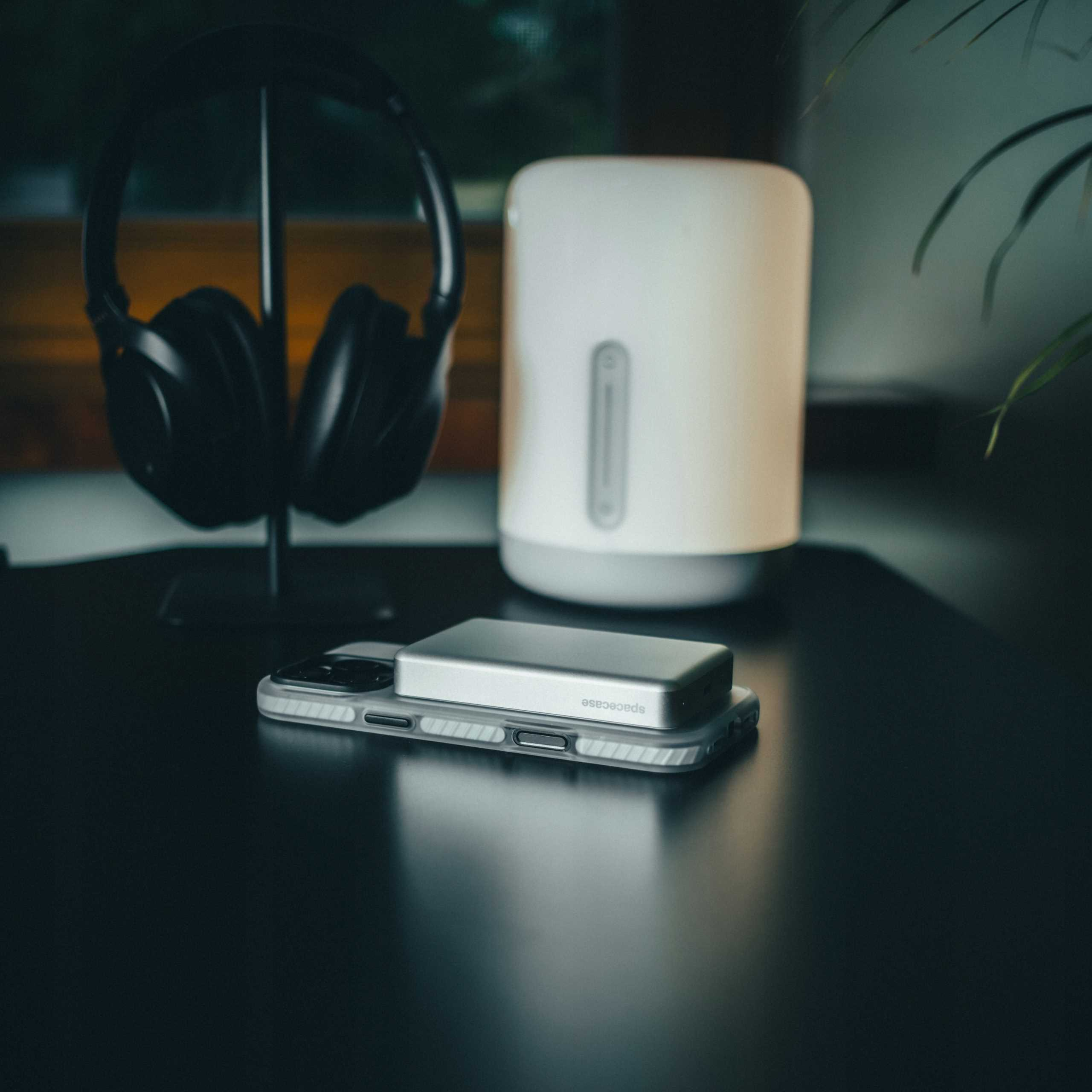 Para czarnych słuchawek na stojaku, białe urządzenie i telefon ze srebrnym akcesorium na ciemnej powierzchni.
