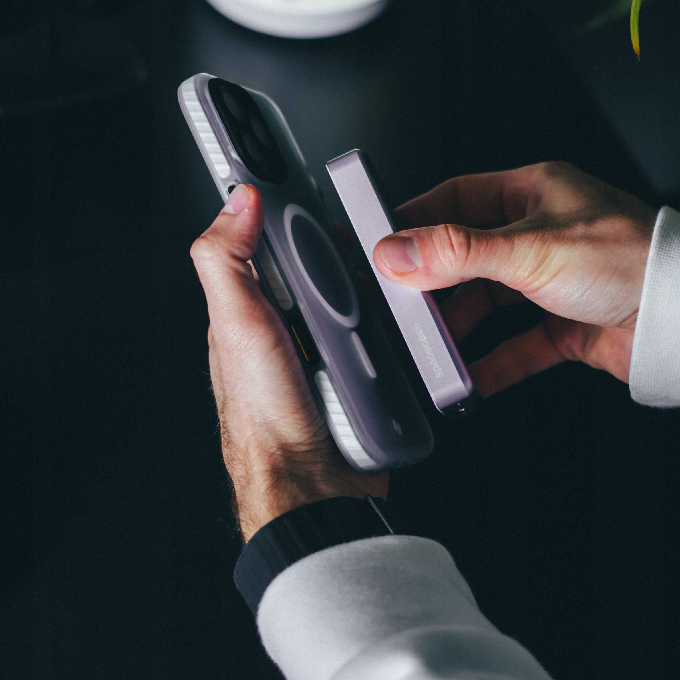 Ręce trzymają telefon i power bank. Telefon ma fioletowe etui. Ciemne tło.