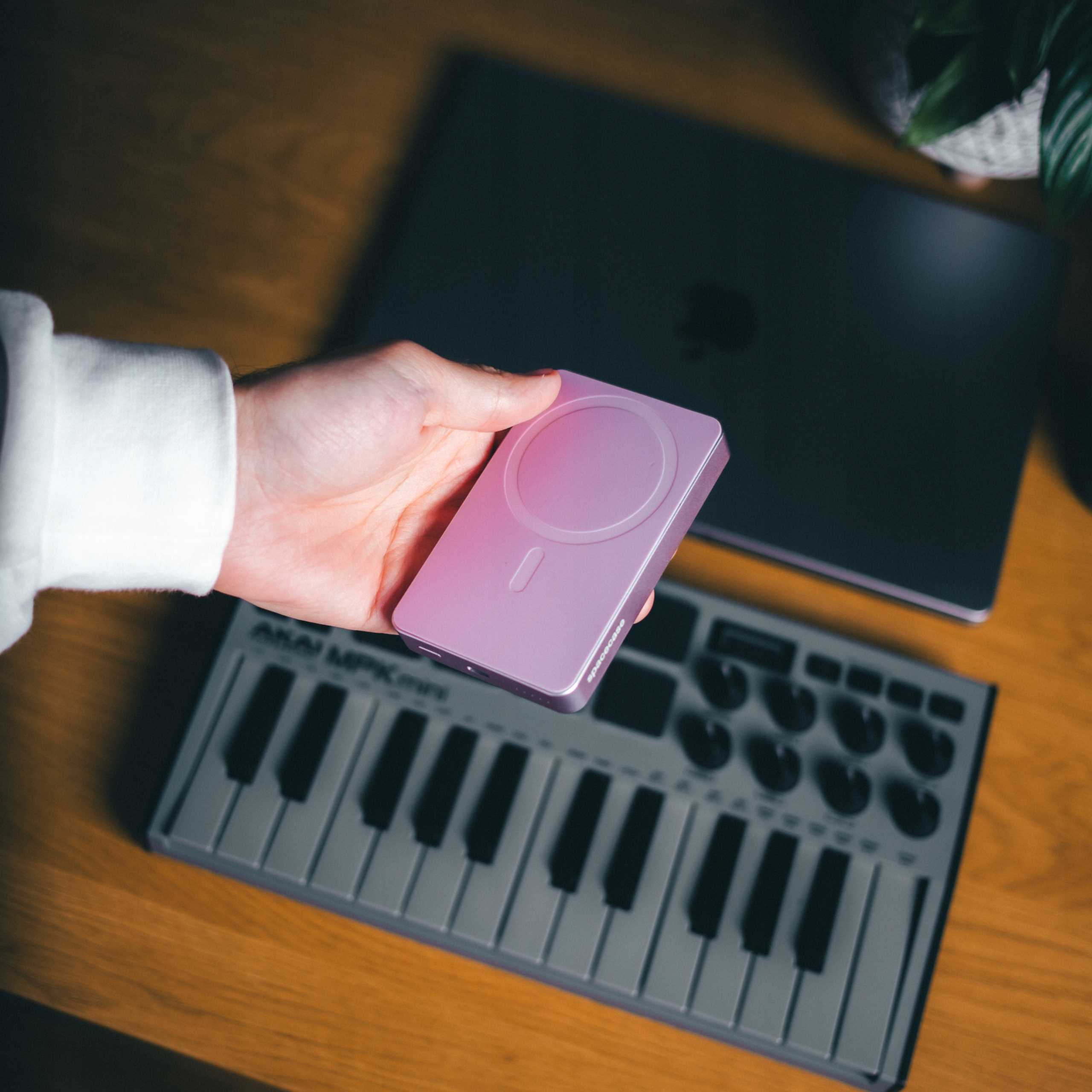 Dłoń trzyma fioletową ładowarkę. Laptop i klawiatura są w tle, na drewnianym stole.