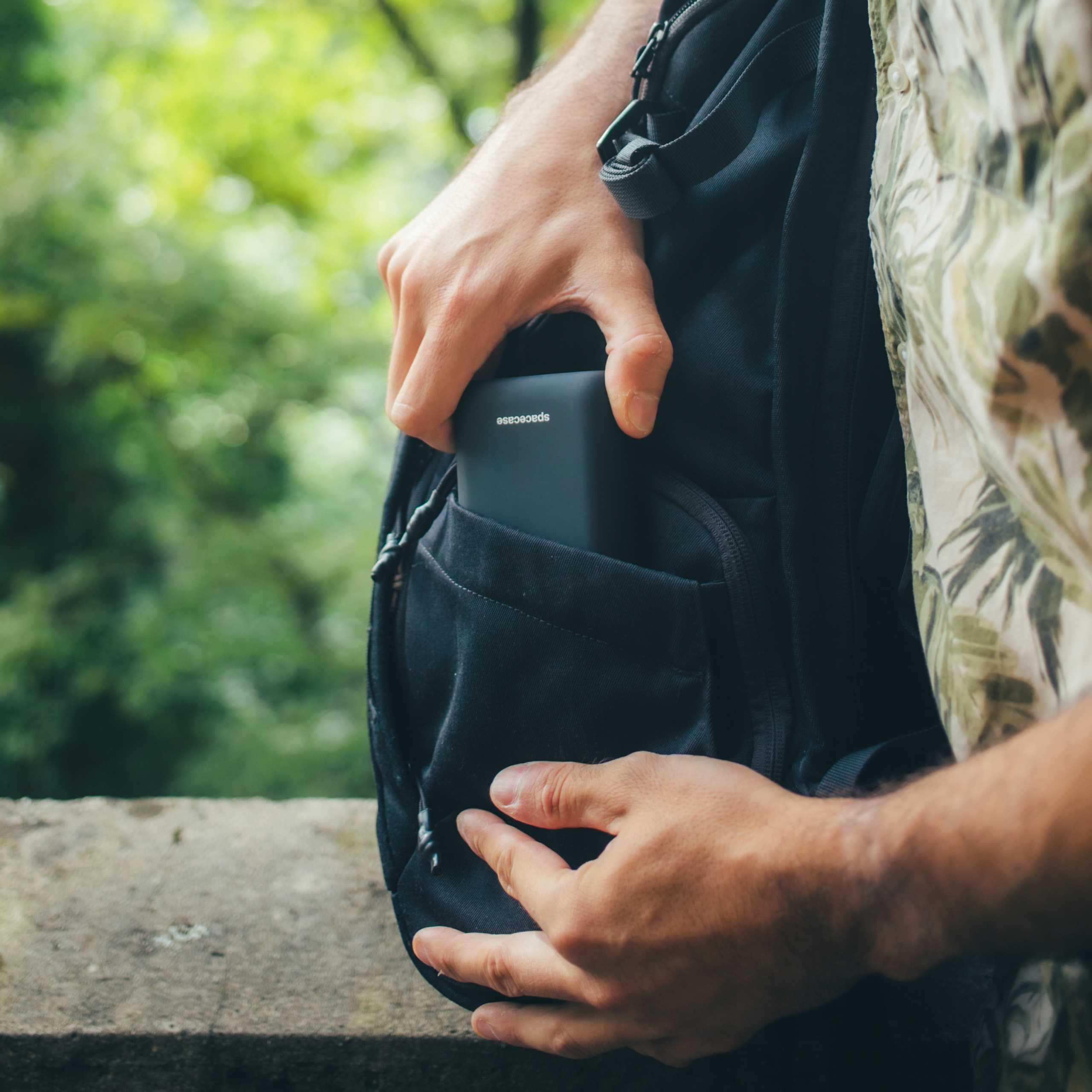 Ręce osoby wkładają czarny power bank spacecase do czarnej kieszeni plecaka. Zielone liście w tle.