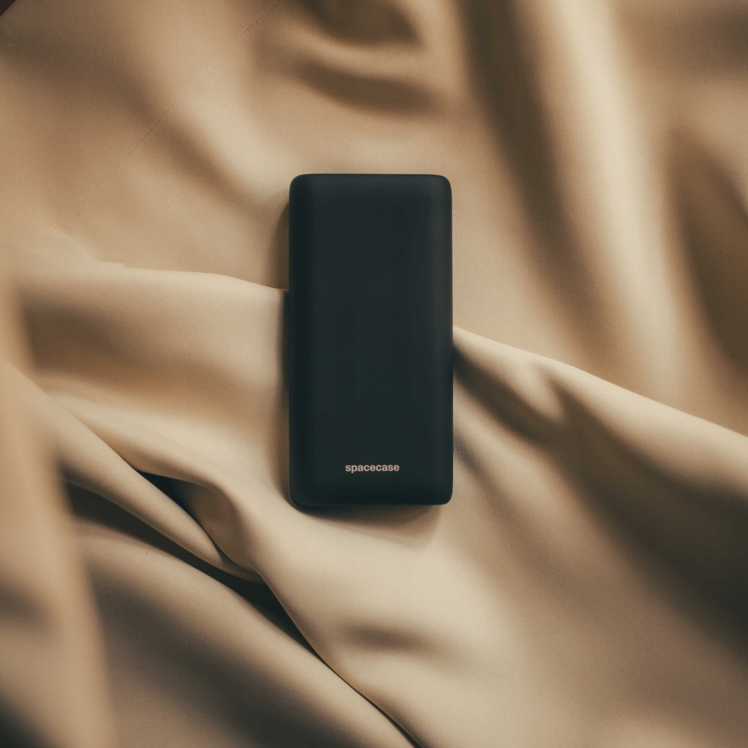 Czarny power bank Spacecase leży na beżowej tkaninie. Tkanina jest miękko udrapowana i teksturowana.