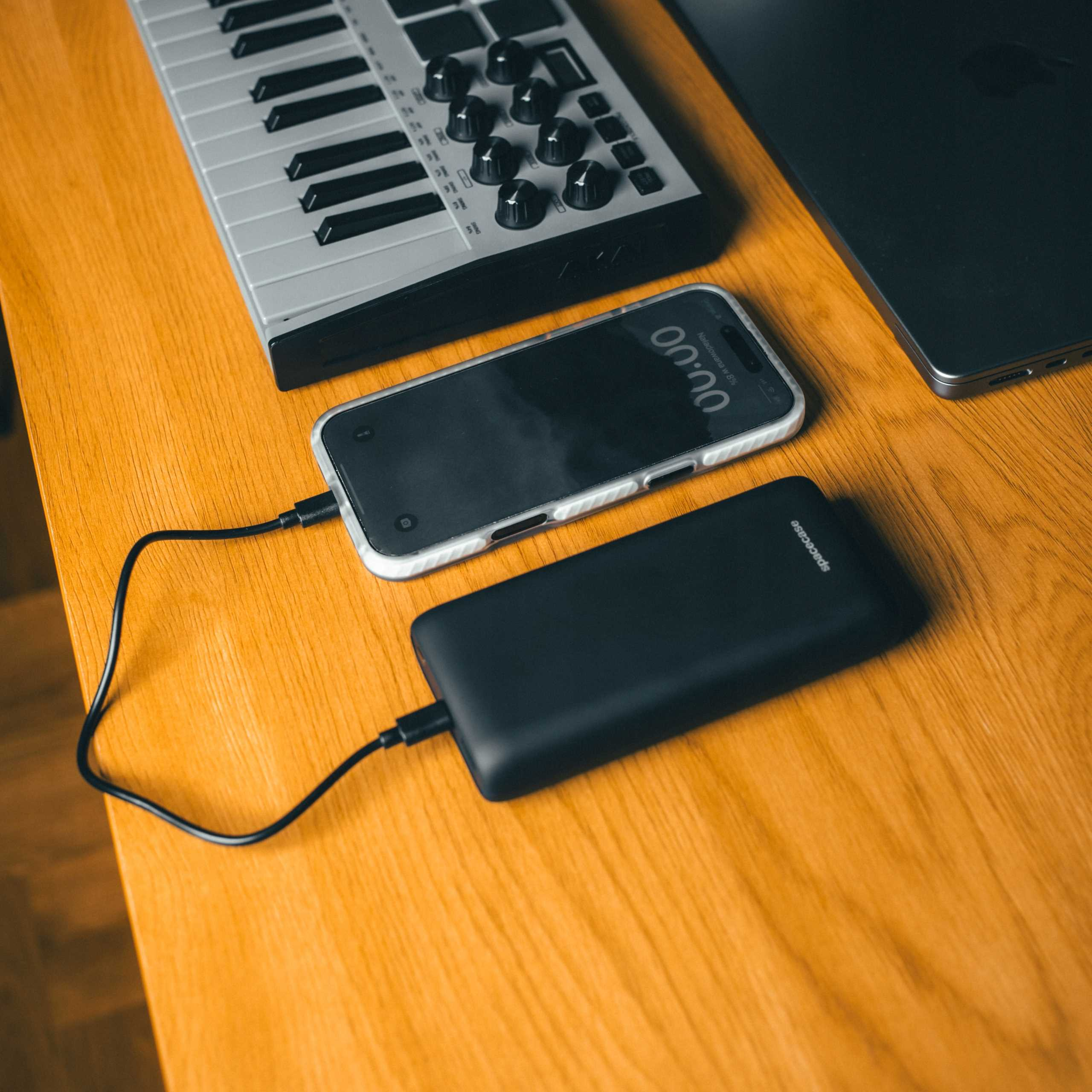 Klawiatura, telefon i power bank leżą na drewnianej powierzchni, połączone kablami ładującymi.