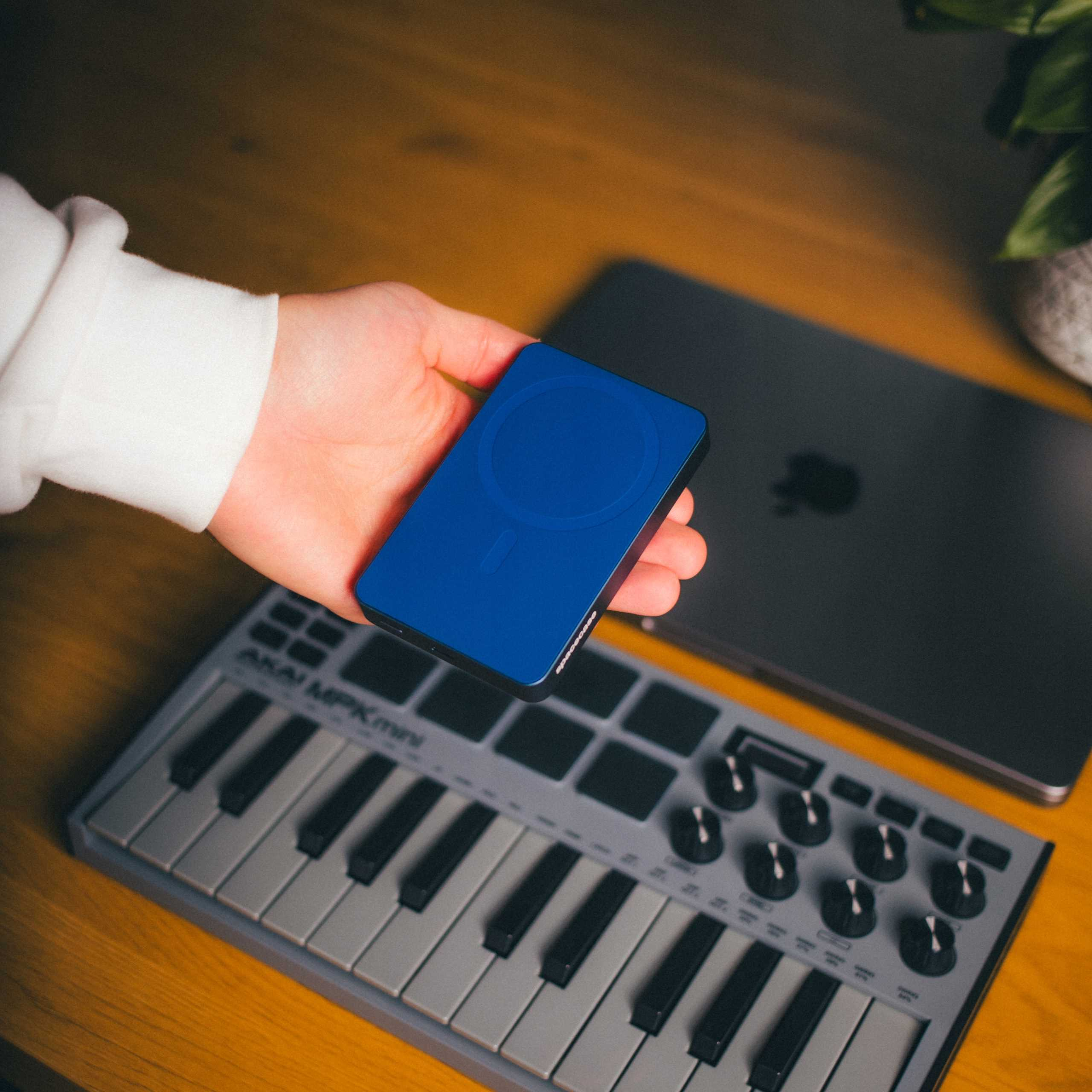 Dłoń trzyma niebieskie prostokątne urządzenie. W tle klawiatura i laptop.