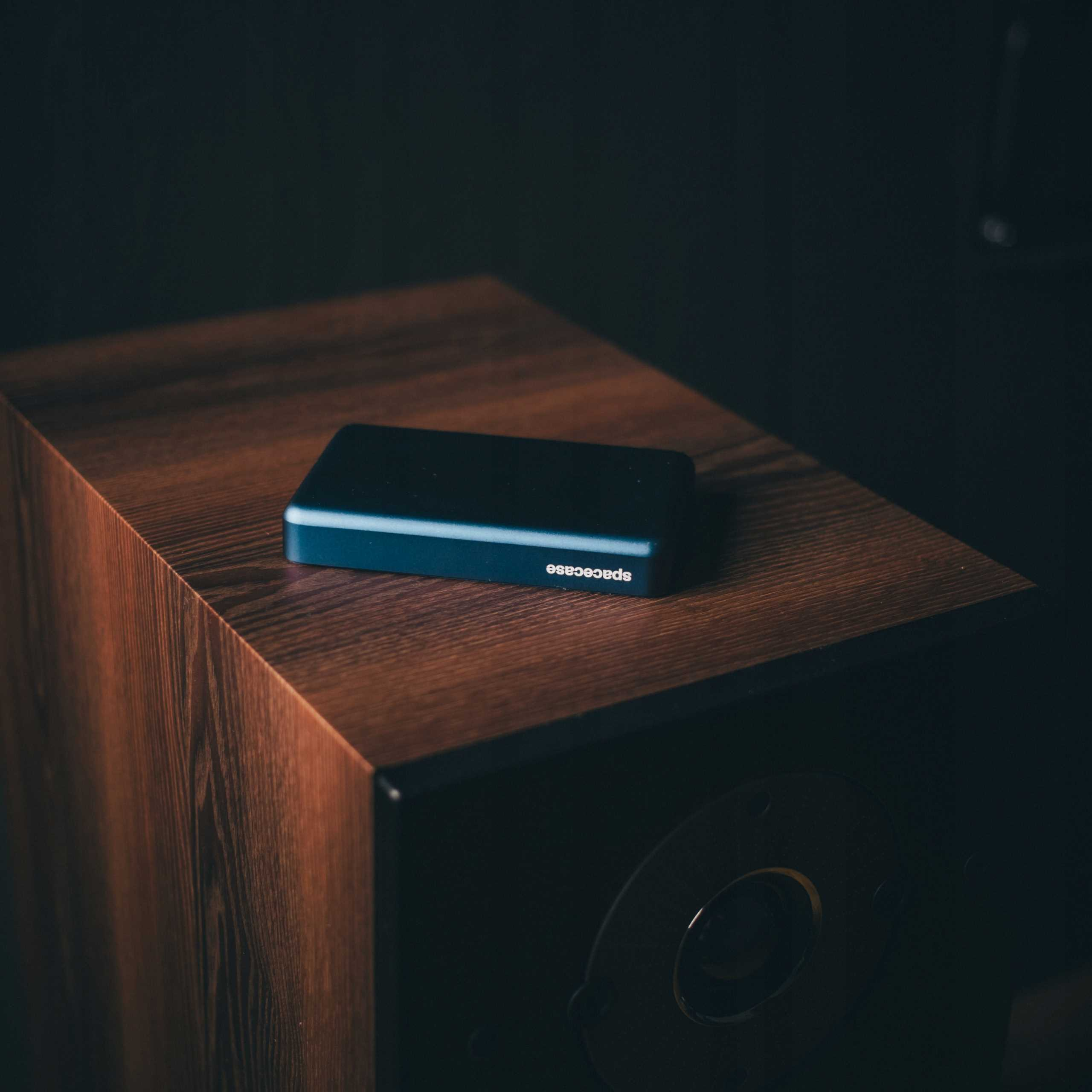 Niebieskie prostokątne urządzenie z napisem 'spacecase' leży na drewnianej skrzyni głośnikowej.