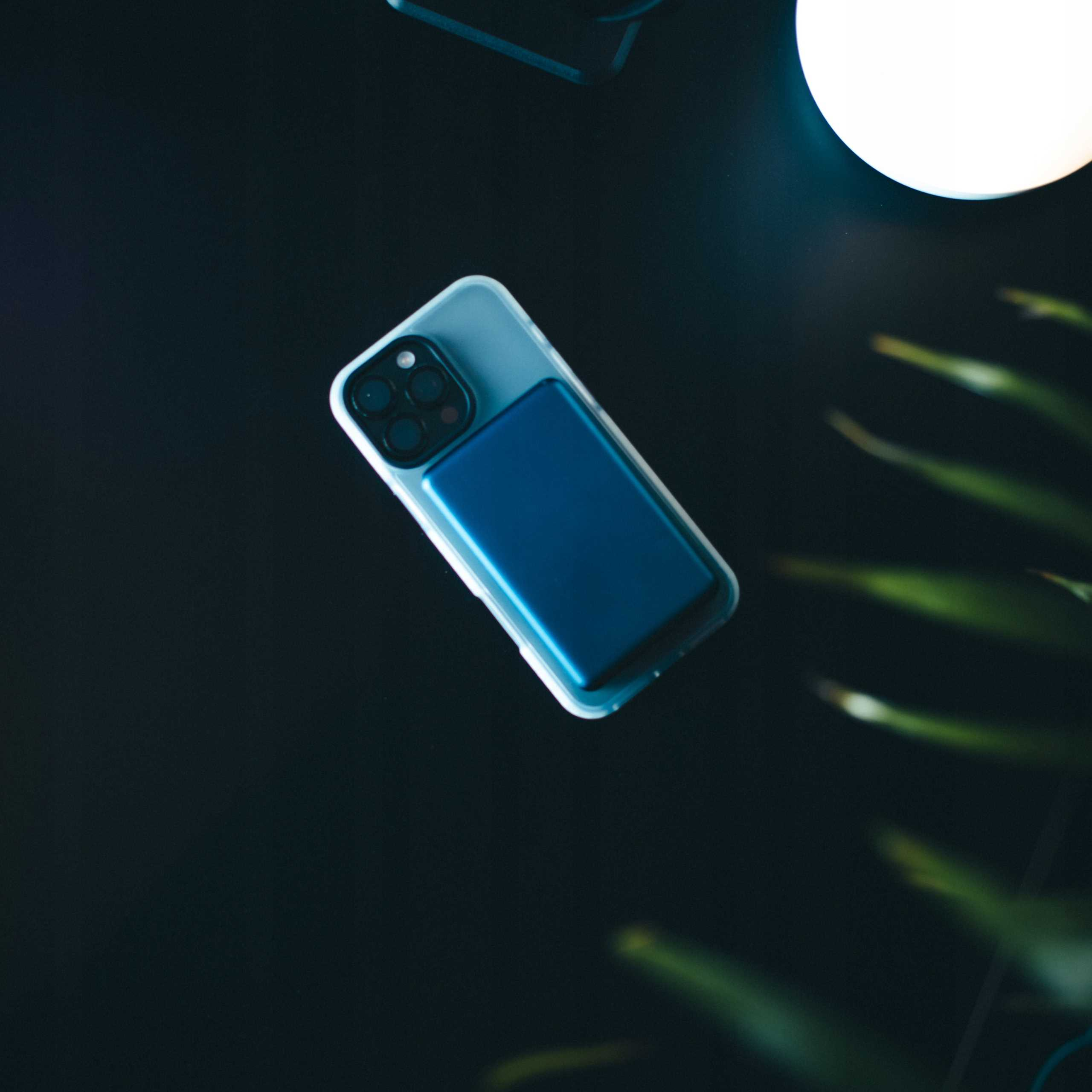 Telefon z niebieską ładowarką na ciemnej powierzchni. Jasne światło i zielone rośliny w tle.