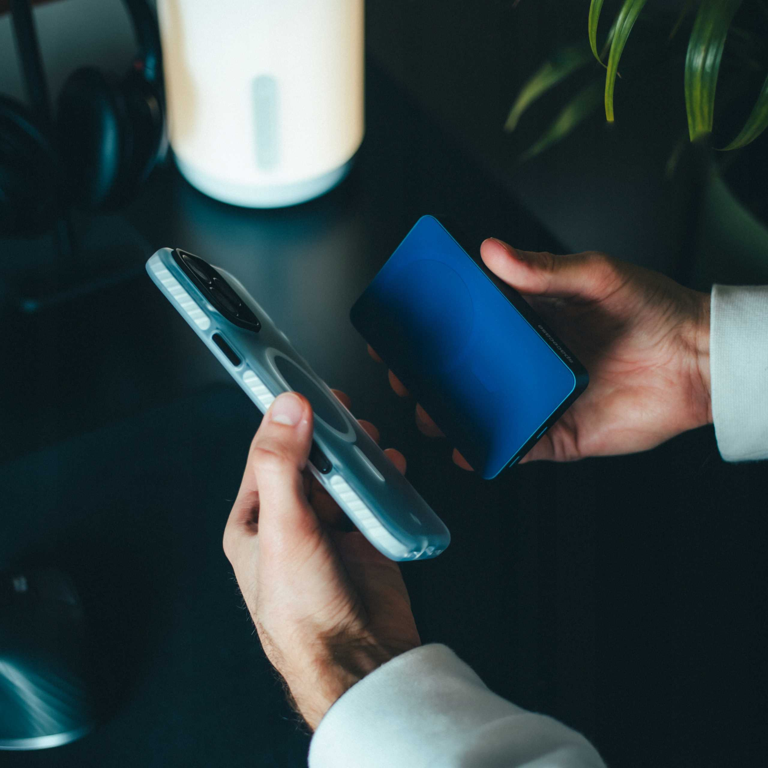 Dłonie trzymające telefon w etui i niebieski power bank. W tle lampa i rośliny.