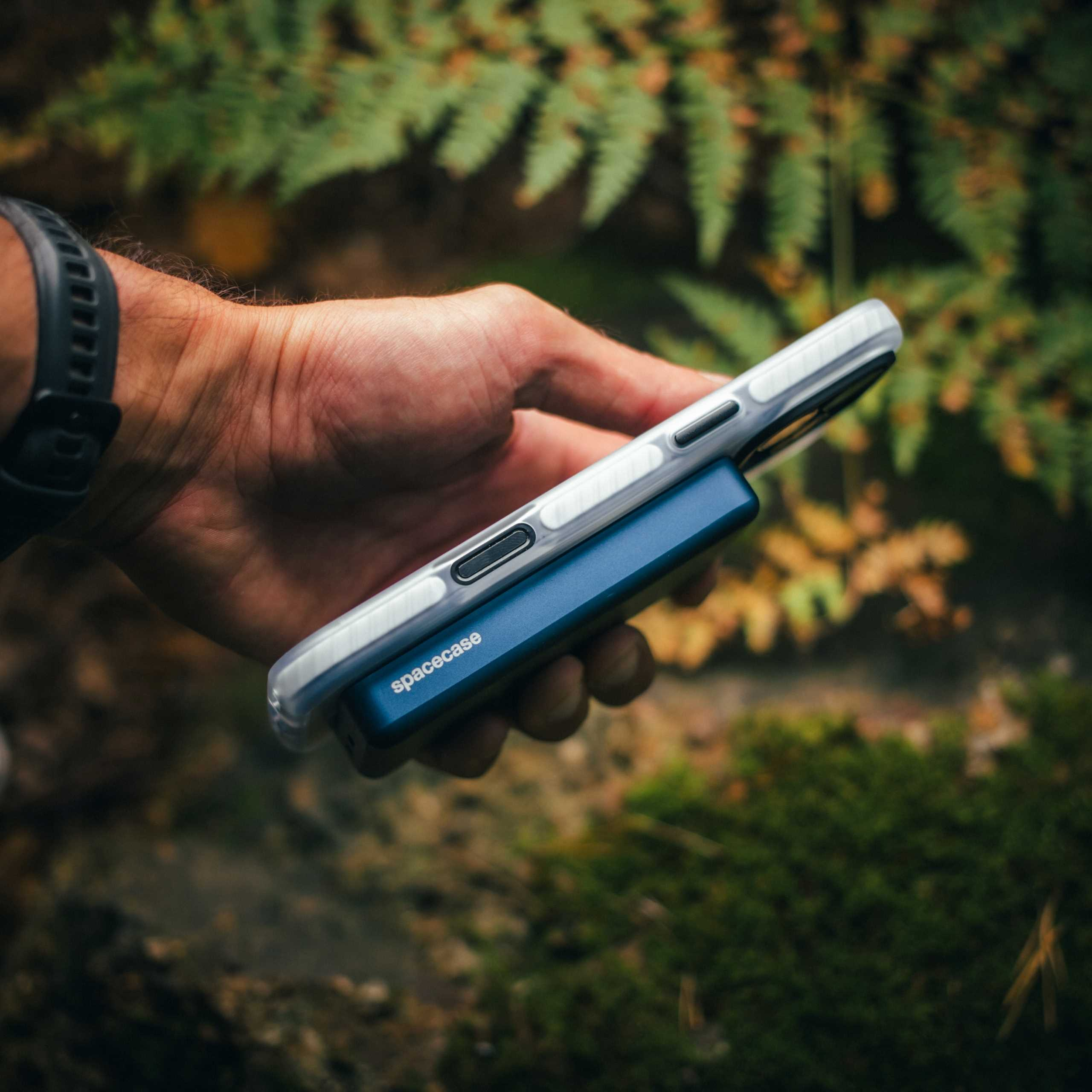 Dłoń trzymająca niebieski power bank Spacecase z telefonem w przezroczystym etui. Zielone liście w tle.