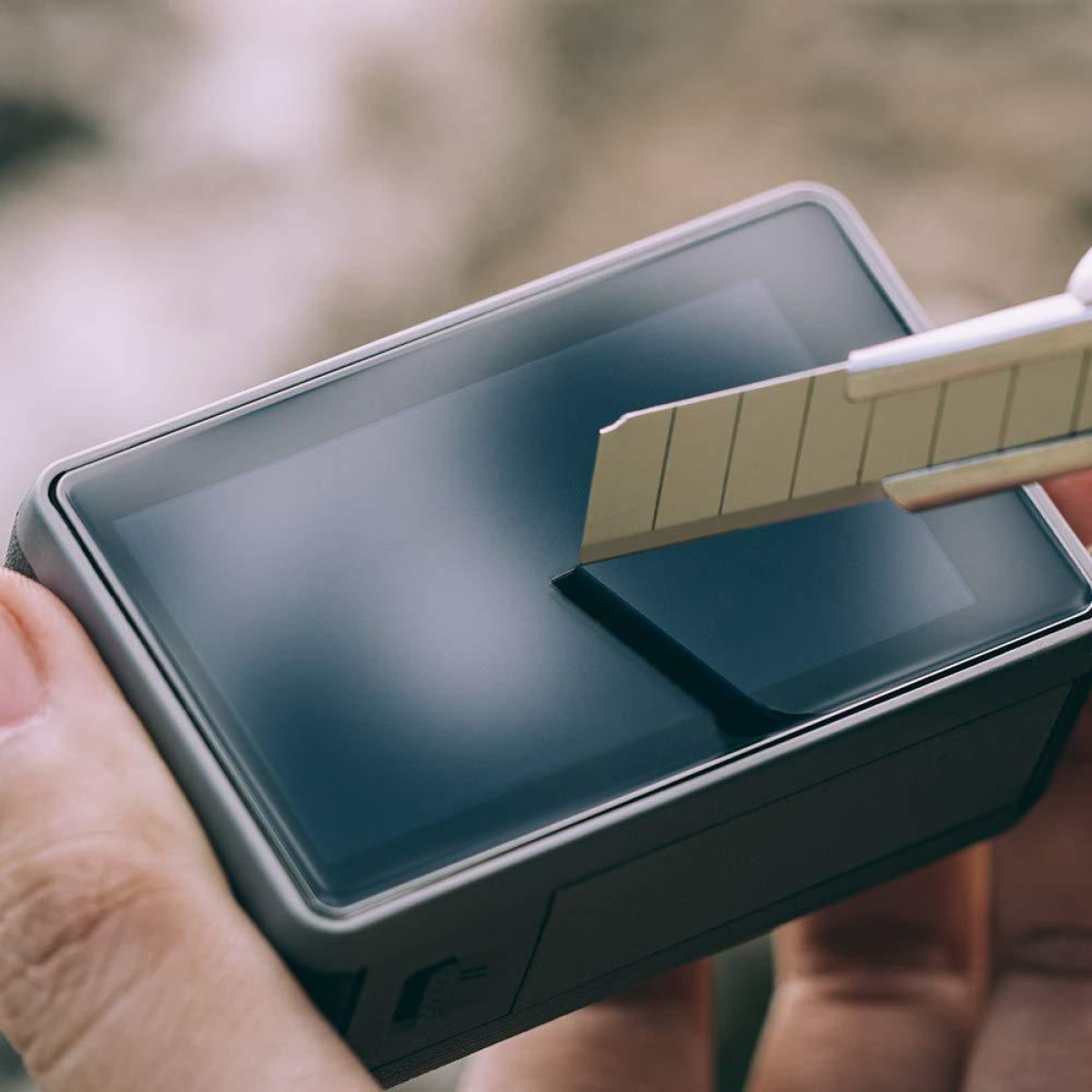 Ręka trzyma urządzenie z ekranem. Ostrze tnie folię ochronną ekranu. Tło jest rozmyte.