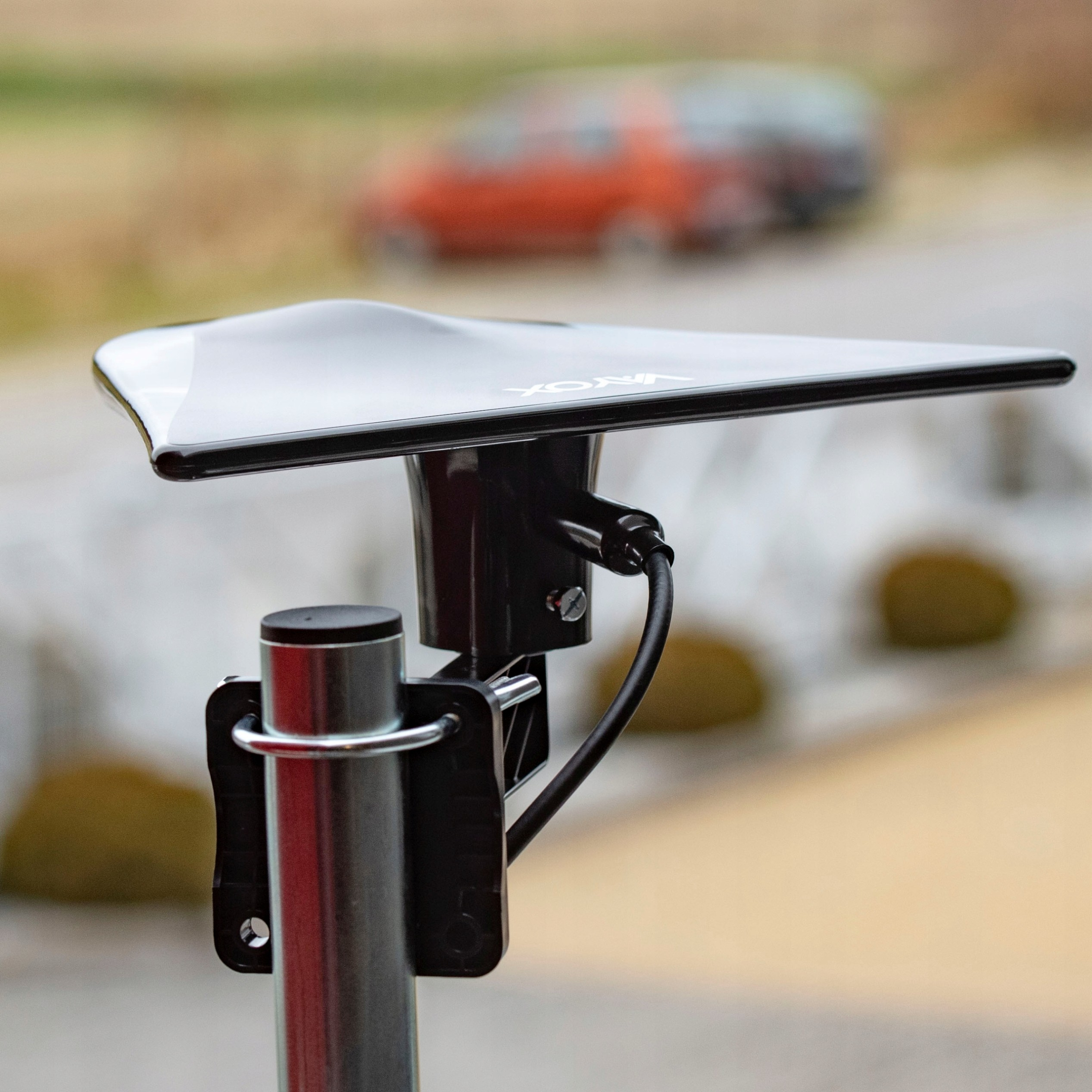 Czarna antena jest zamontowana na słupie. Pomarańczowy samochód jest rozmyty w tle. Na antenie napis "Voxin".