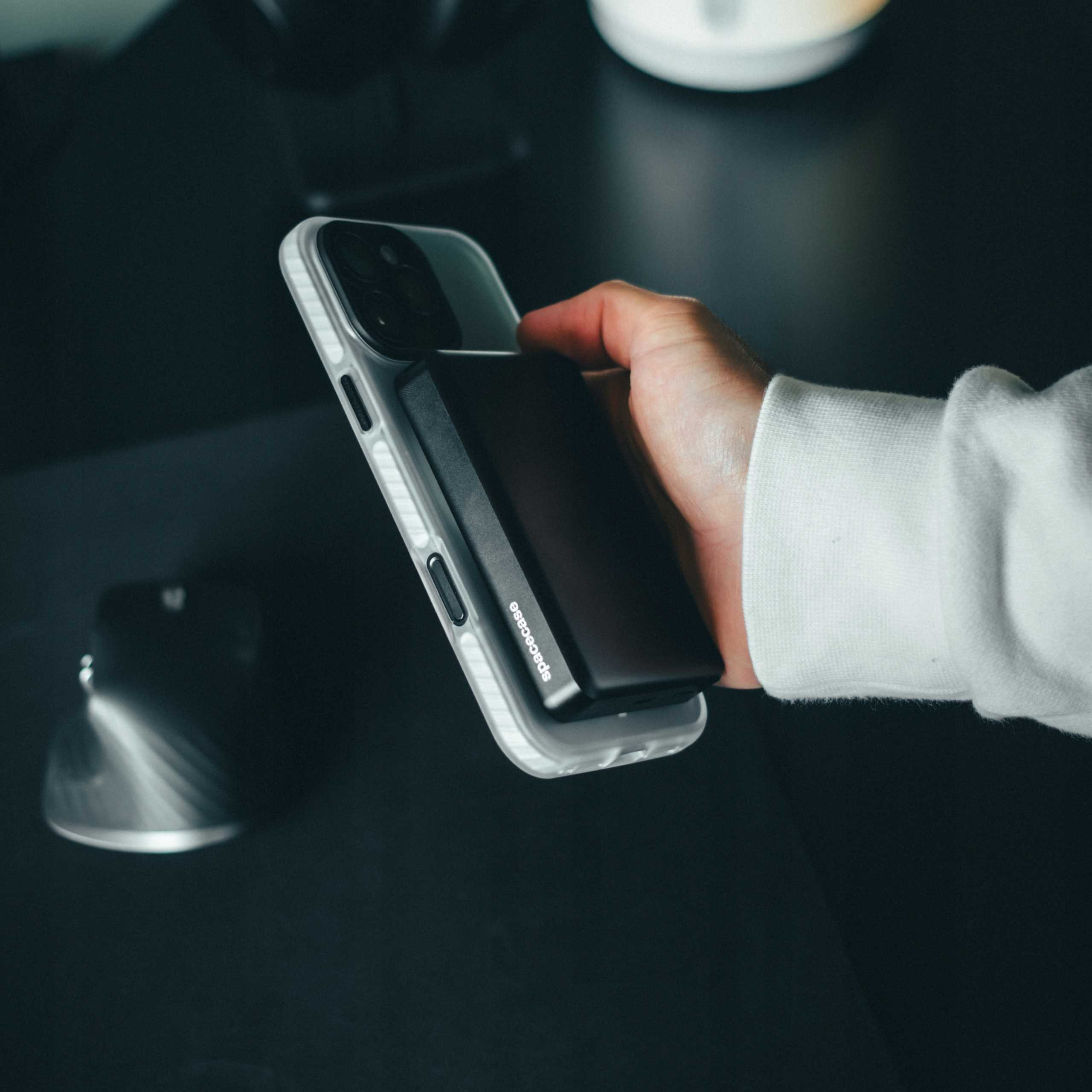 Dłoń trzymająca telefon z czarnym akcesorium. Widoczny biały rękaw. Ciemne tło.