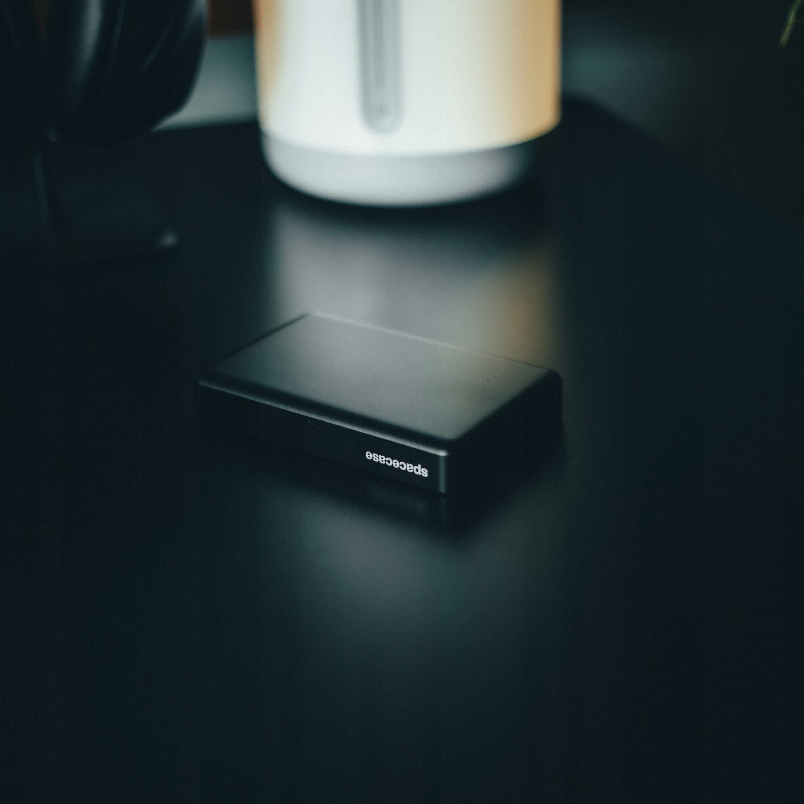 Czarny przenośny dysk twardy z napisem 'spacecase' leży na odblaskowej czarnej powierzchni. Biały czajnik jest rozmyty w tle.