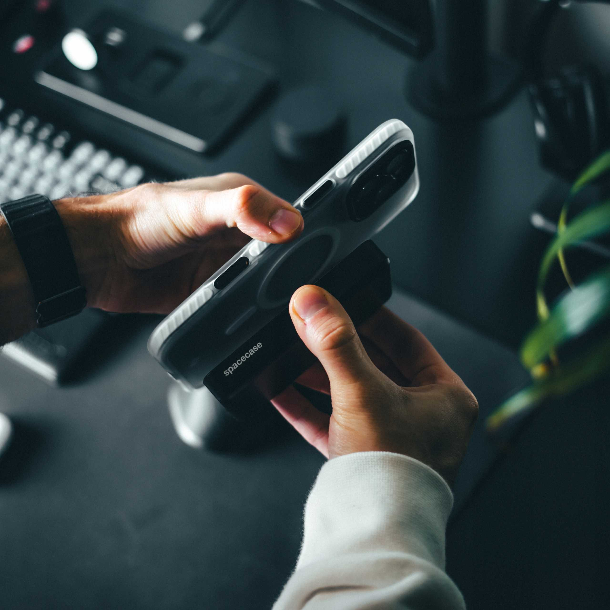 Ręce trzymają telefon w przezroczystym etui na czarnej powierzchni. Zegarek widoczny na lewym nadgarstku.