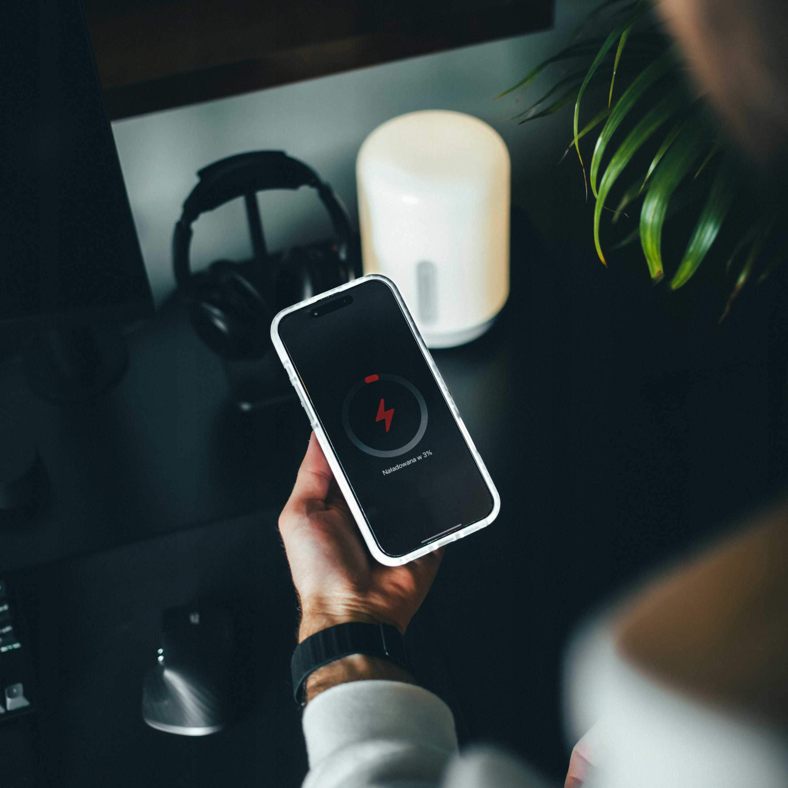 Ręka trzyma telefon, który się ładuje. Czerwona błyskawica na ekranie. Inne przedmioty na biurku.