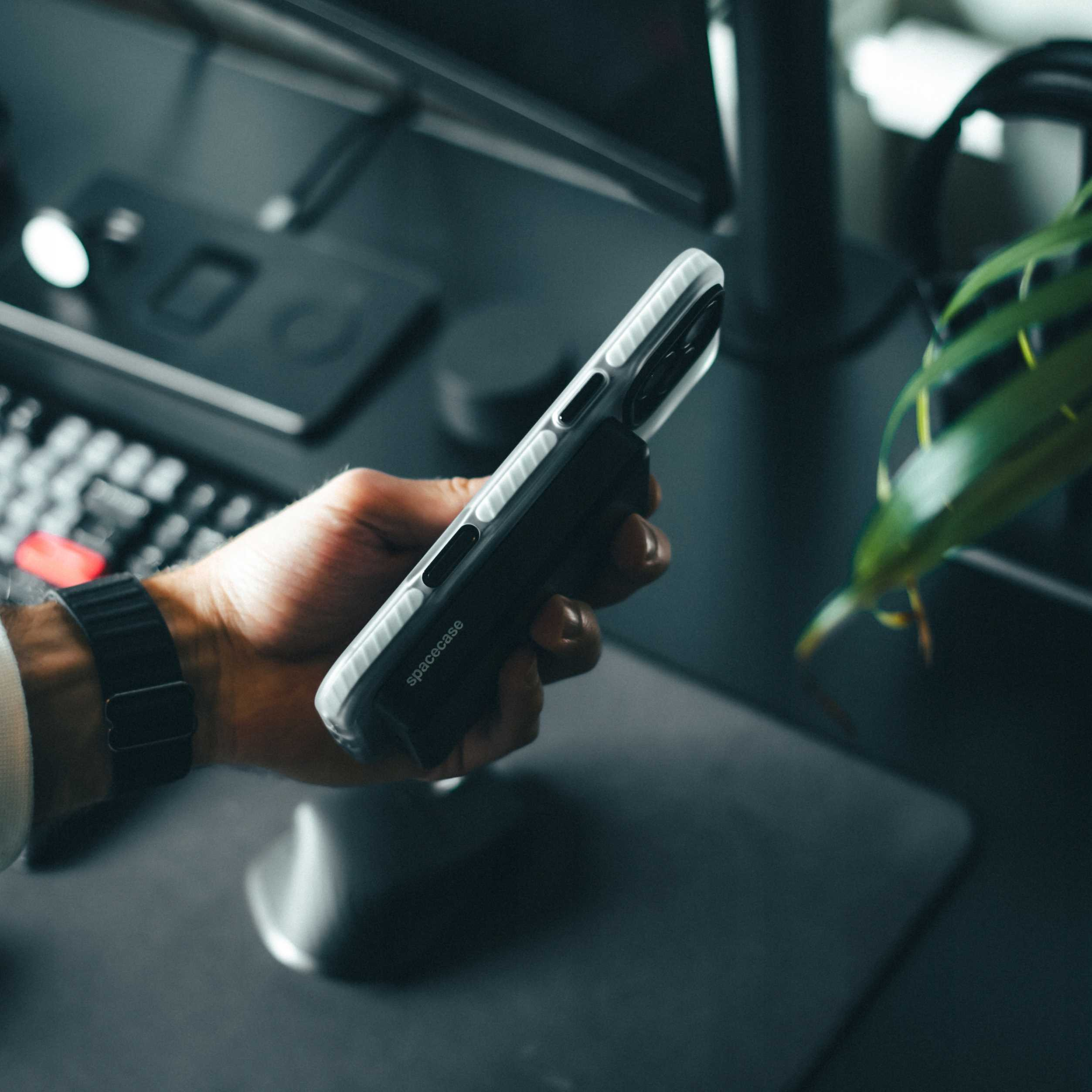 Dłoń trzyma telefon w etui spacecase. Klawiatura, podkładka pod mysz i roślina w rozmytym tle.