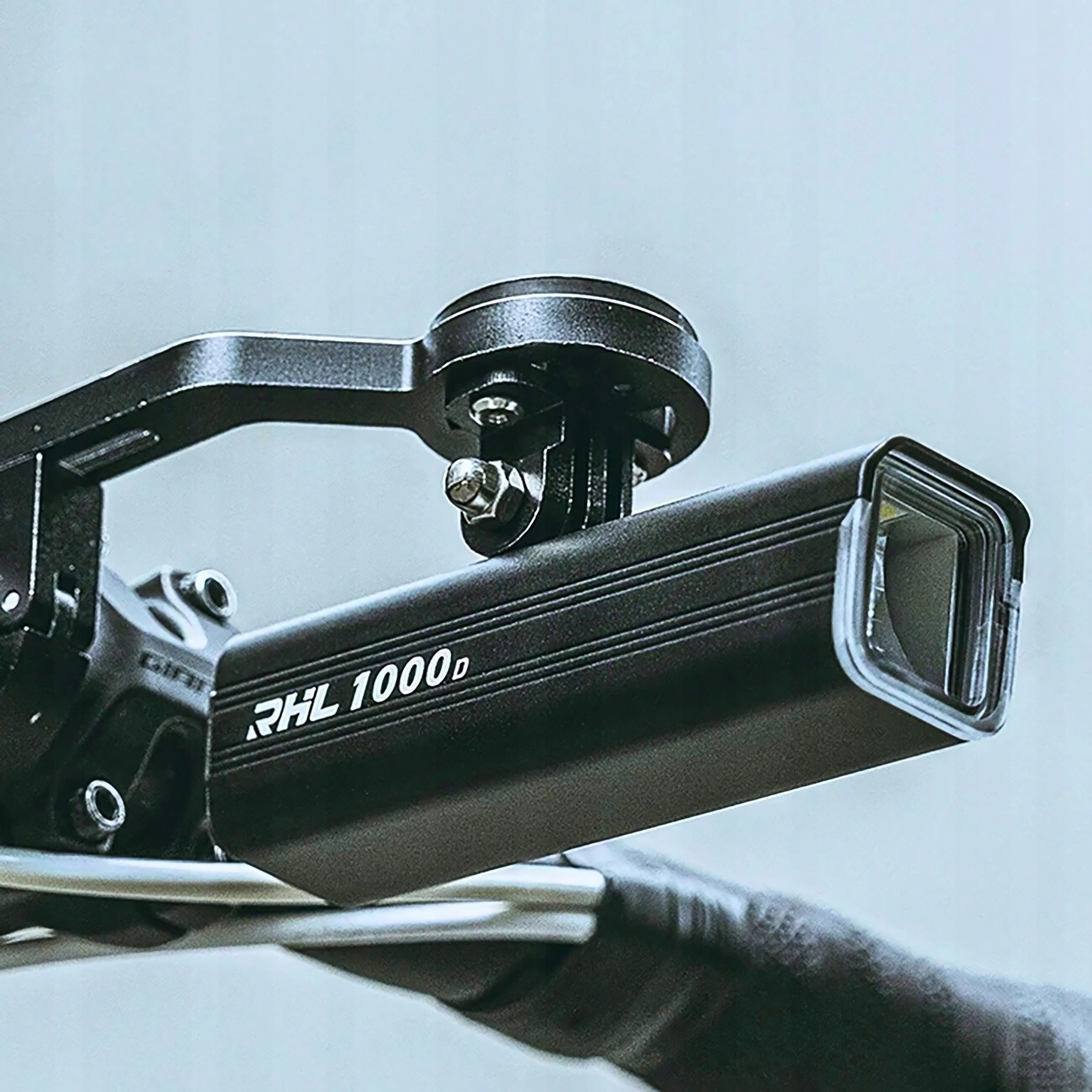 Zbliżenie czarnej lampy rowerowej zamontowanej na kierownicy. Oznaczenie RHL 1000d. Tło jest rozmyte.