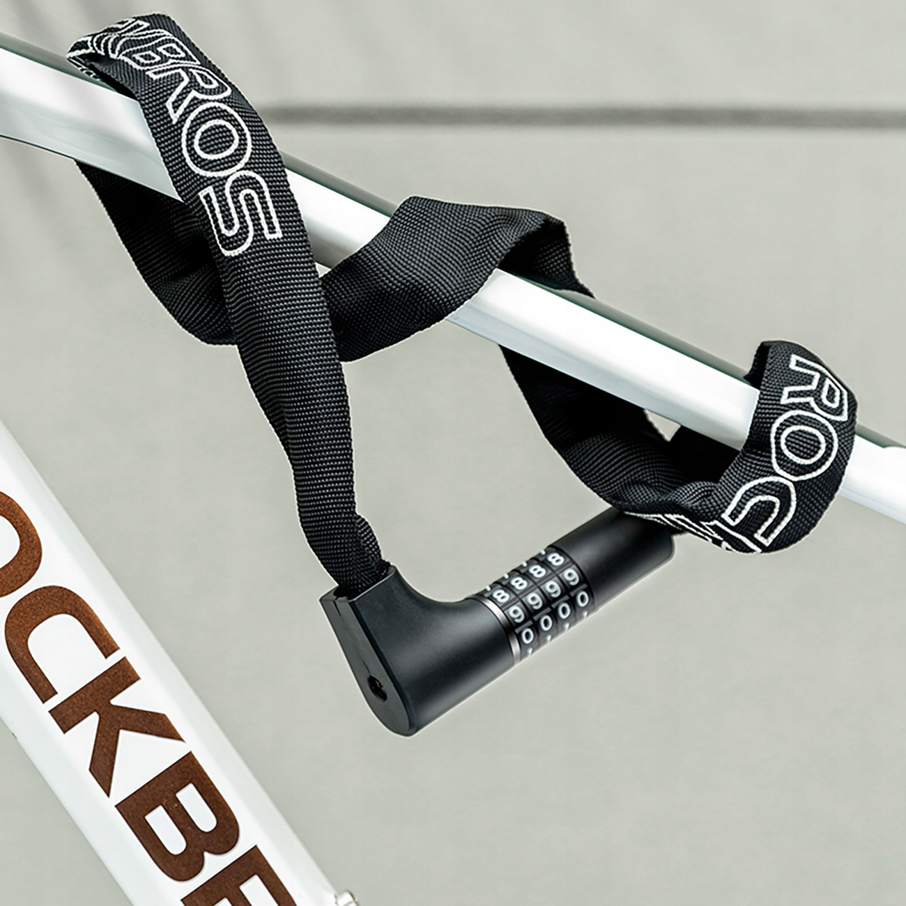 Czarna blokada rowerowa jest owinięta wokół białej ramy roweru. Zamek ma zamek szyfrowy i nazwę marki.