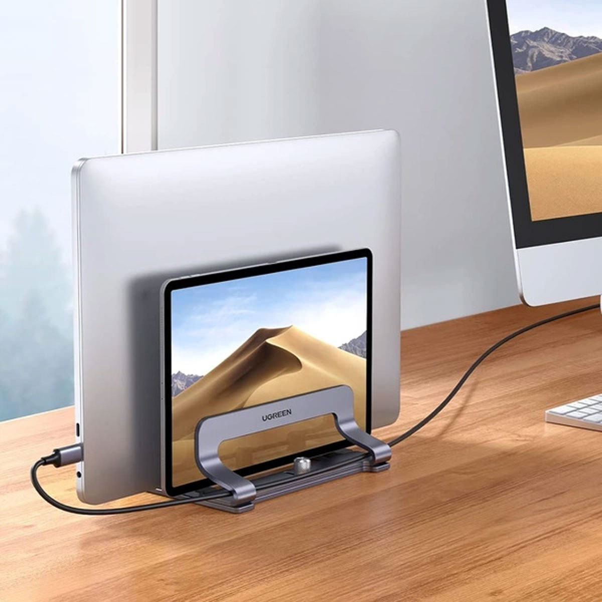 Laptop i tablet są trzymane przez srebrny uchwyt na drewnianym biurku. Ekran tabletu pokazuje pustynny krajobraz.