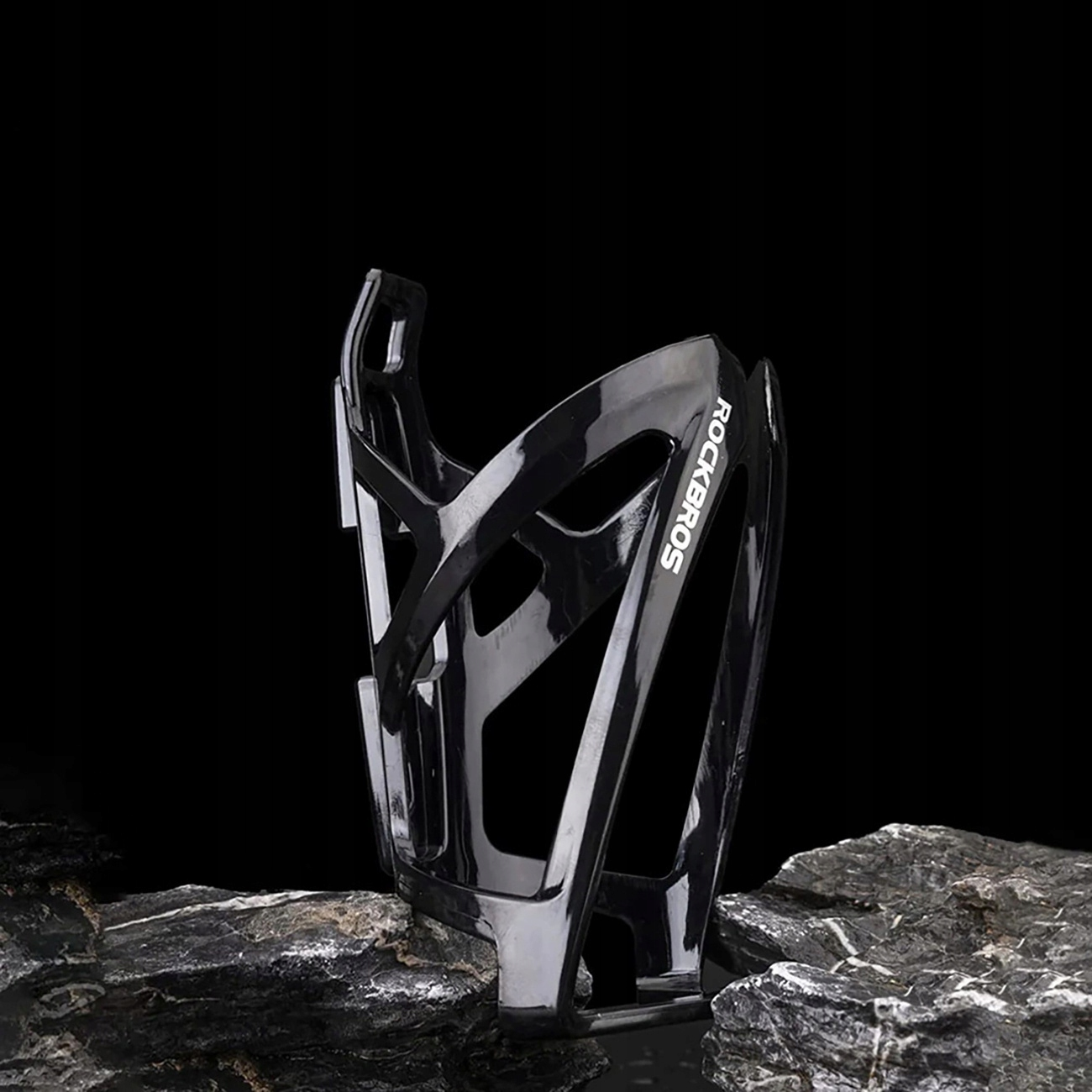 Czarny koszyk na bidon Rockbros. Stoi na ciemnej, skalistej powierzchni na czarnym tle. Koszyk jest pusty.