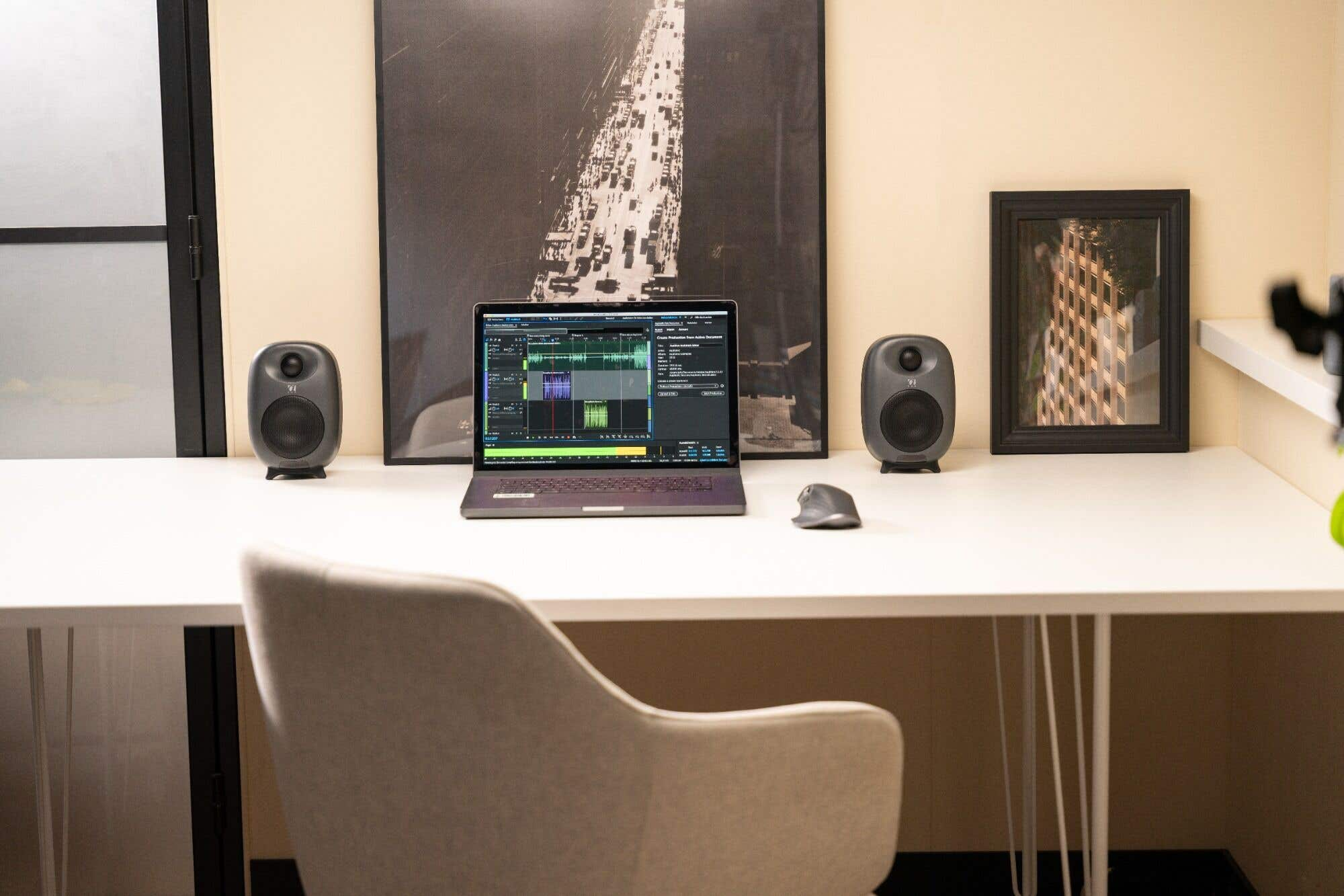 Un espacio de trabajo con una laptop mostrando software de edición de audio, altavoces, un ratón y fotos enmarcadas.