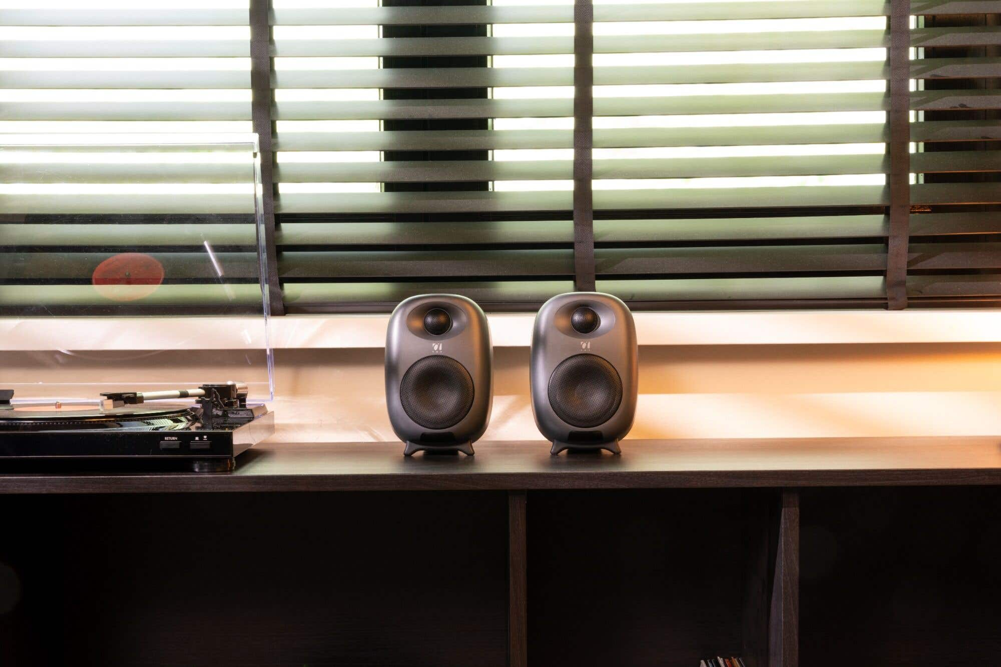 Dos altavoces grises están en un estante oscuro con un tocadiscos. Las persianas están en el fondo.