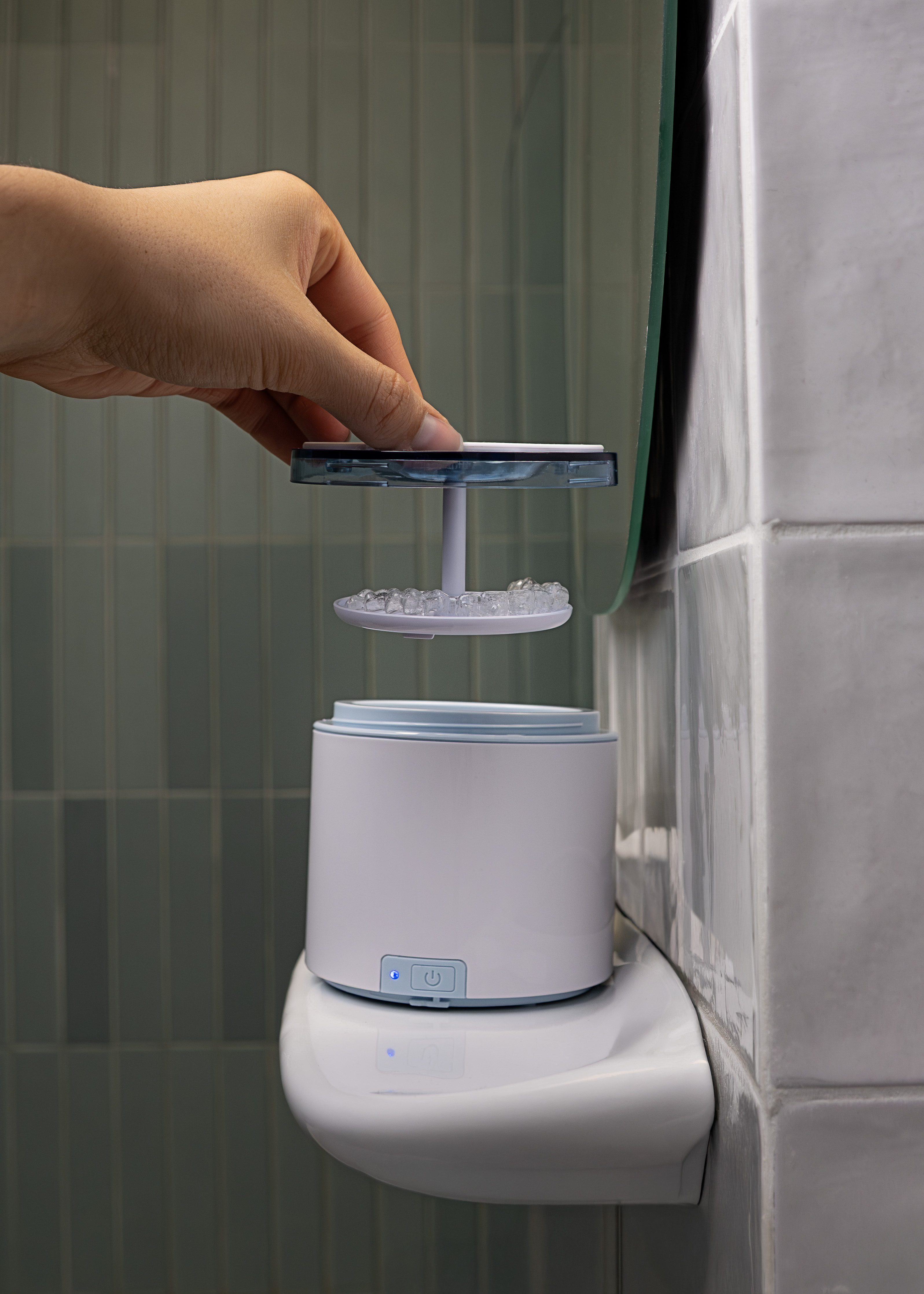 Ręka umieszcza aligner na urządzeniu. Urządzenie jest białe, cylindryczne i ma przycisk. Ściana jest wyłożona kafelkami.