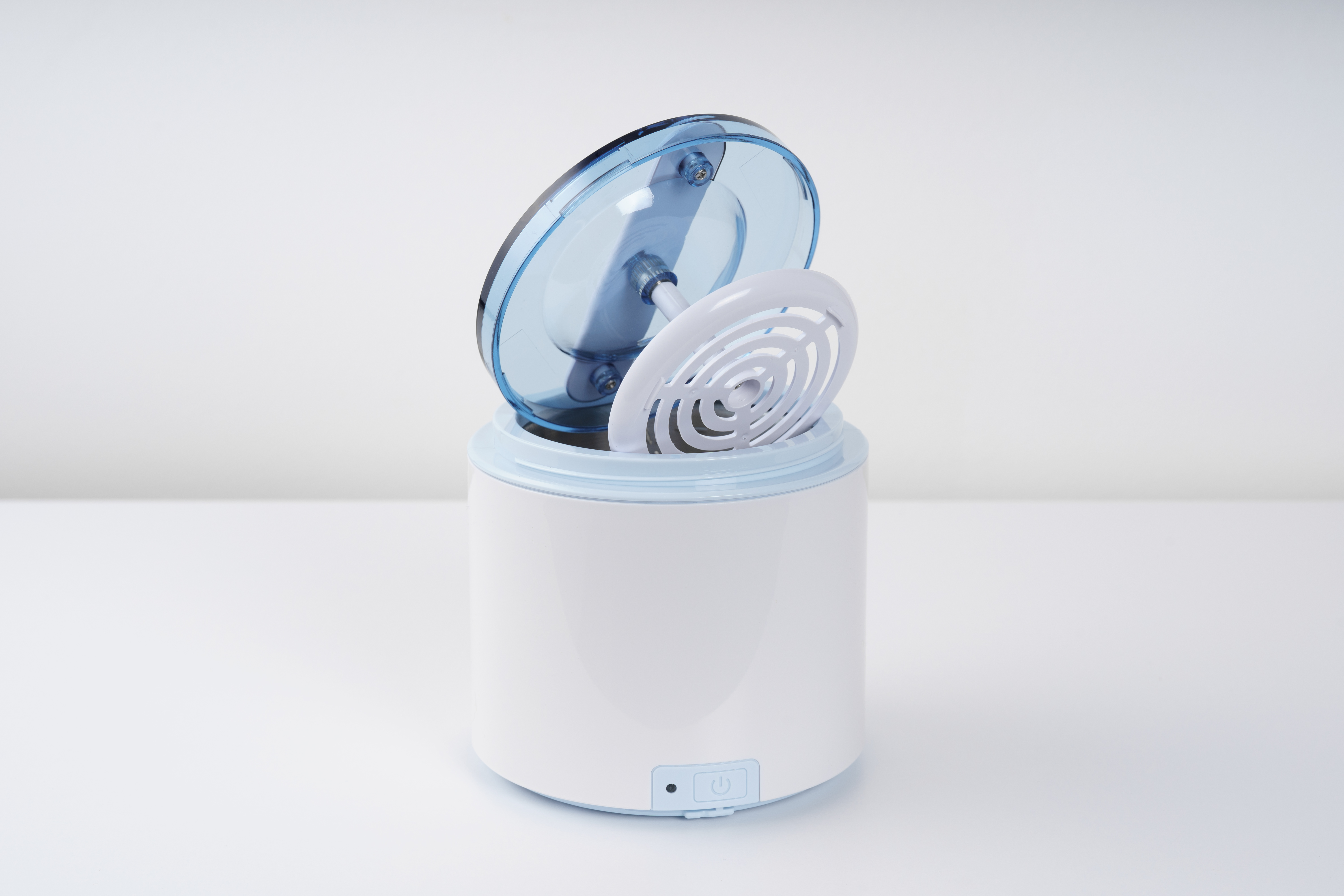 Białe i niebieskie urządzenie z otwartą pokrywą, odsłaniające białą okrągłą część w środku. Stoi na białej powierzchni.