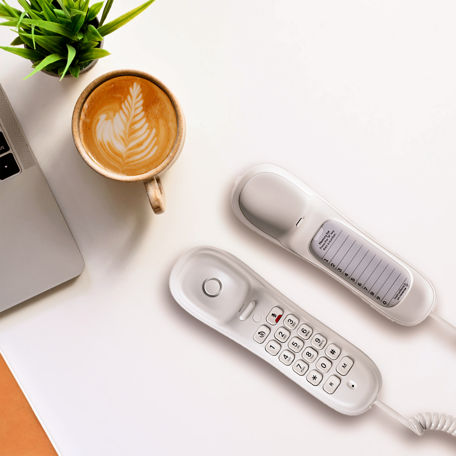 Een bovenaanzicht van een wit bureau met een laptop, een koffiekopje met latte art, een kleine plant en twee witte telefoons.