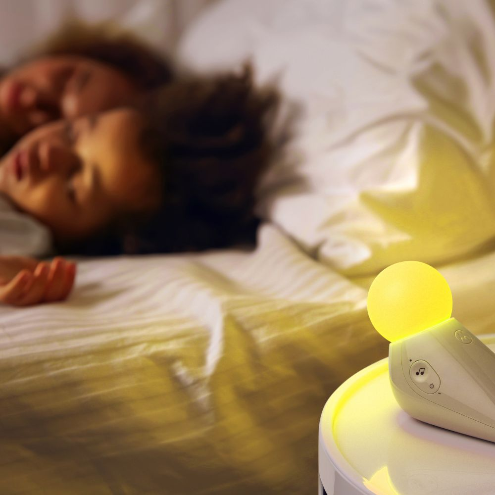 Zwei Personen schlafen in einem Bett. Ein sanftes gelbes Nachtlicht leuchtet auf einem Nachttisch. Die Bettwäsche ist weiß.