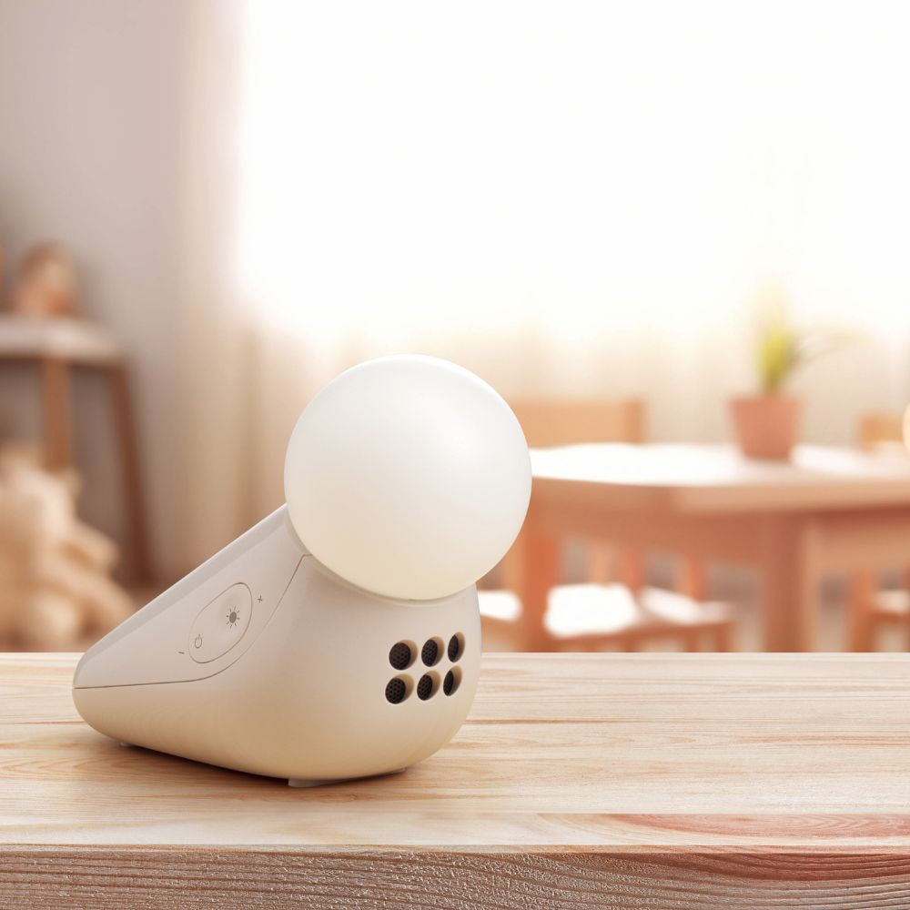 Eine kleine, beige Lampe mit kugelförmigem Licht steht auf einem Holztisch. Der Hintergrund zeigt einen verschwommenen Raum mit Tisch und Pflanze.
