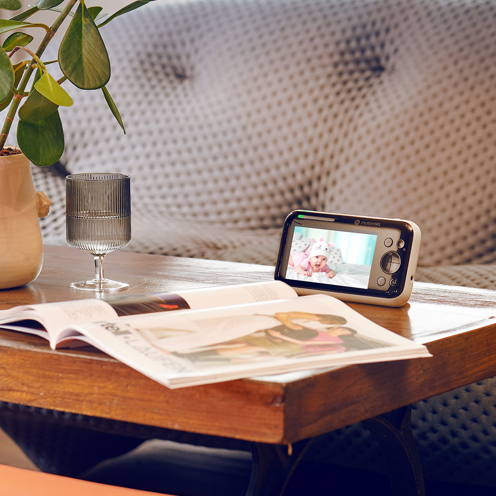 Ein Babyphone auf einem Tisch zeigt ein Baby in Rosa. Ein Glas, ein Magazin und eine Pflanze sind ebenfalls auf dem Holztisch.