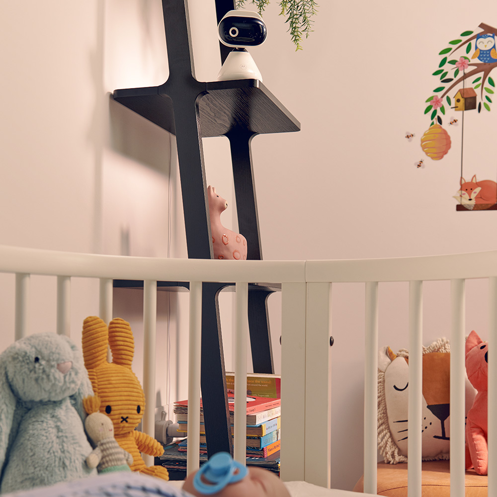 Ein Babybett mit Stofftieren und einem Monitor auf einem Regal. Eine Wanddekoration hat eine Eule, einen Fuchs und einen Bienenkorb.